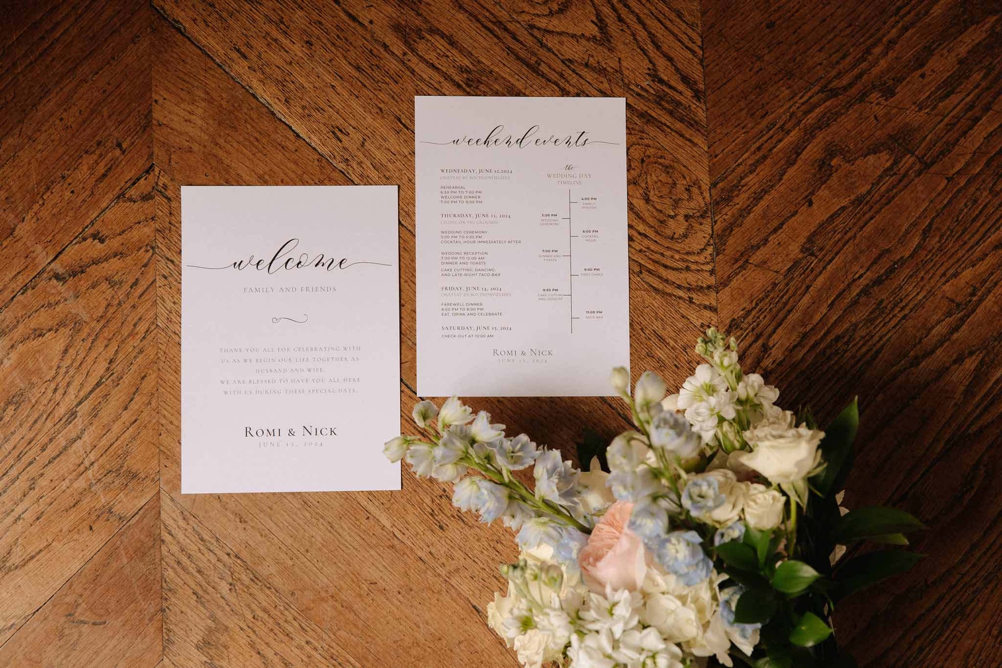 Flat lay of wedding welcome card and weekend schedule on dark wood floor with white stock flowers and blush roses