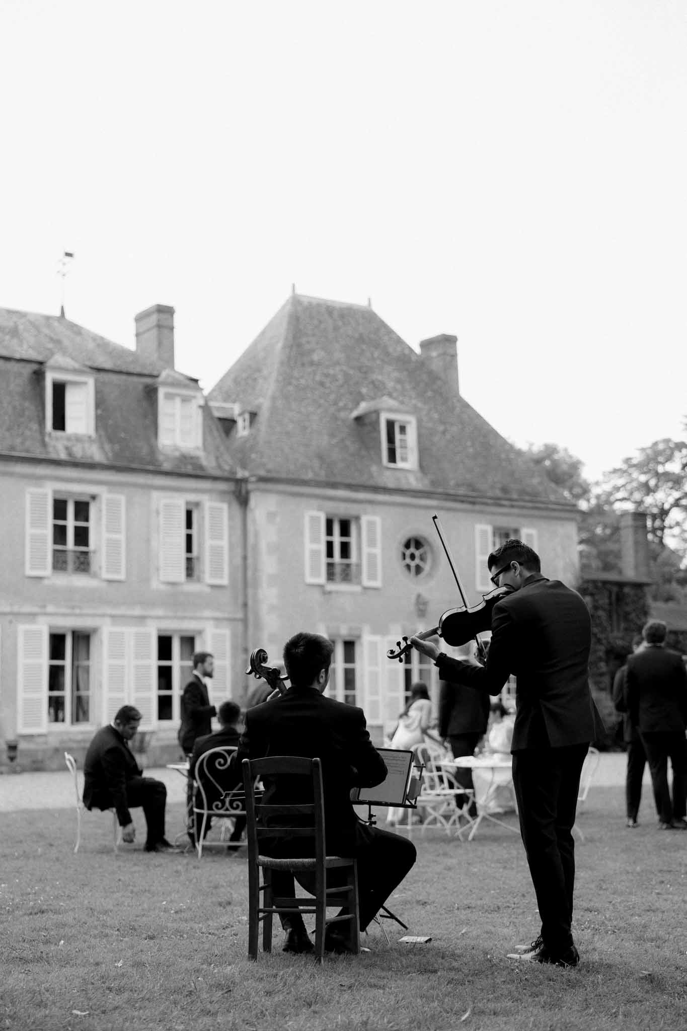 Black and white photo of two musicians playing violin and cello for guests in the courtyard of a French chÃ¢teau