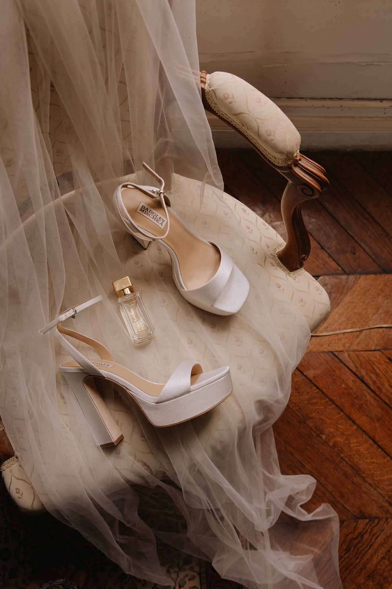 Flat lay of bridal accessories on ivory tulle including two pairs of white platform heels, perfume bottle and gold jewelry