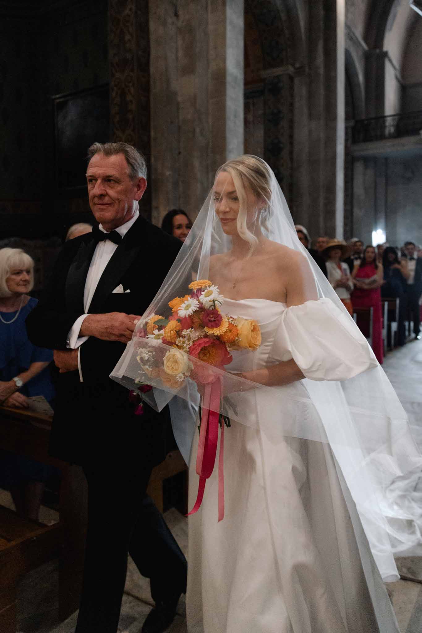 Bride and father walk down the aisle in a stone church with arched columns as guests watch from pews