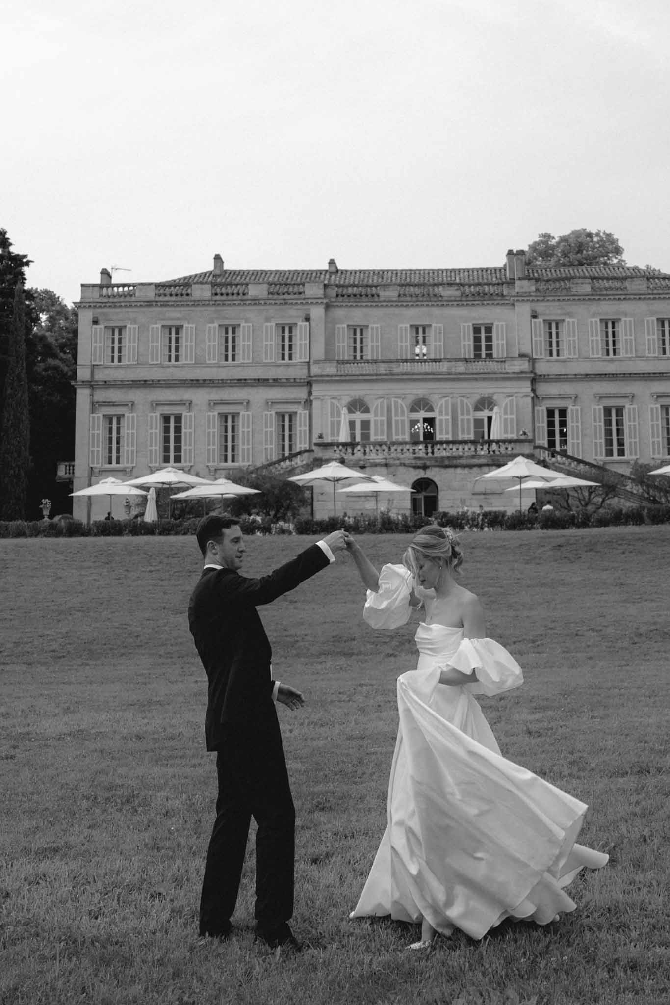 Black and white photo of couple dancing on lawn before a neoclassical chateau with reception umbrellas
