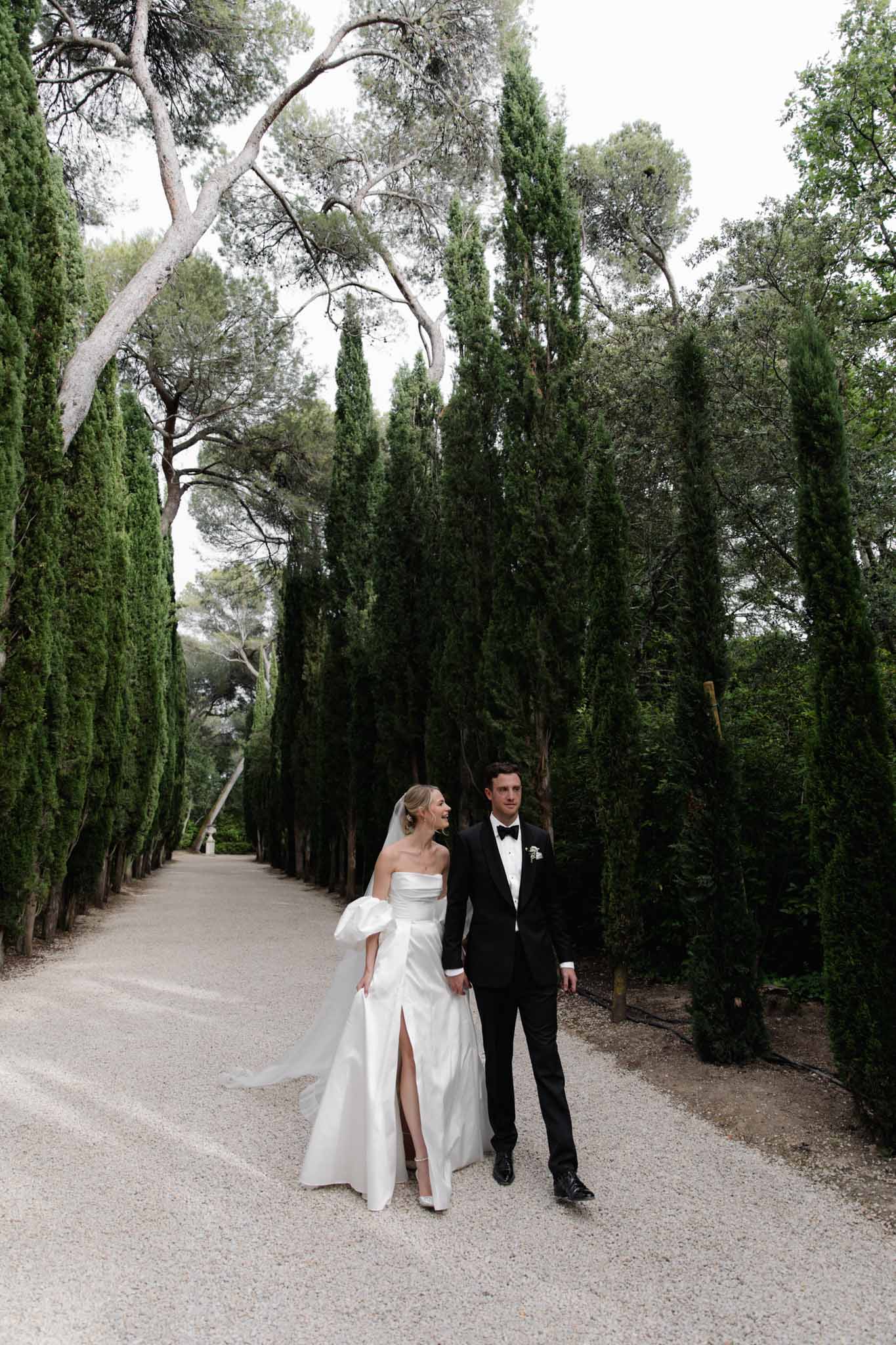 Bride and groom walking down cypress-lined gravel pathway at formal estate with natural tree canopy overhead