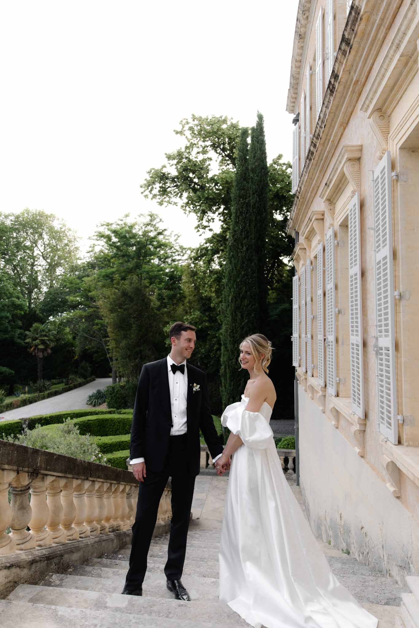 Bride and groom walking hand-in-hand along a stone terrace beside a classical French chateau during portrait session
