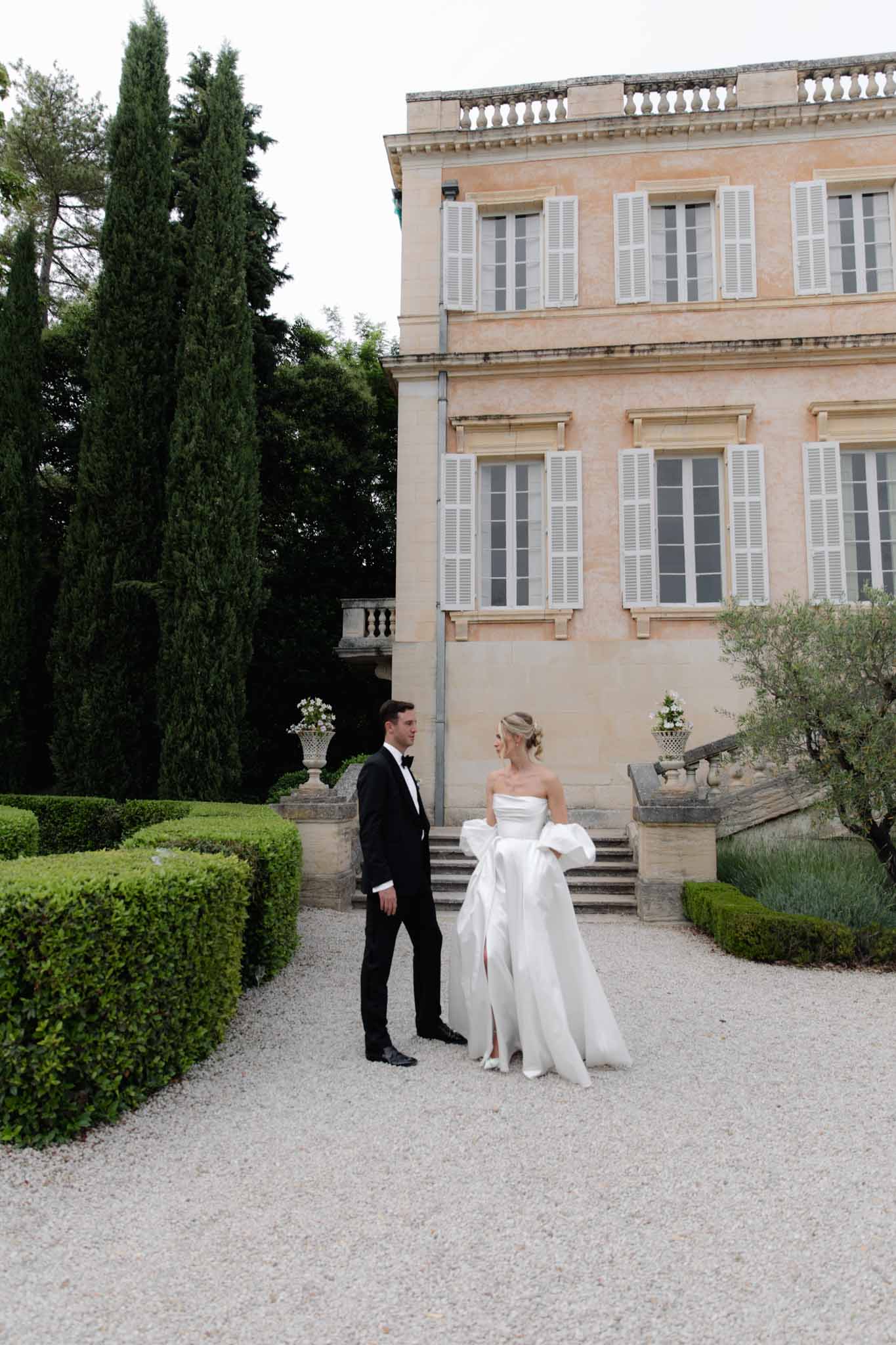 Couple in formal attire on gravel courtyard before pale stone manor with cypress trees and stone urns