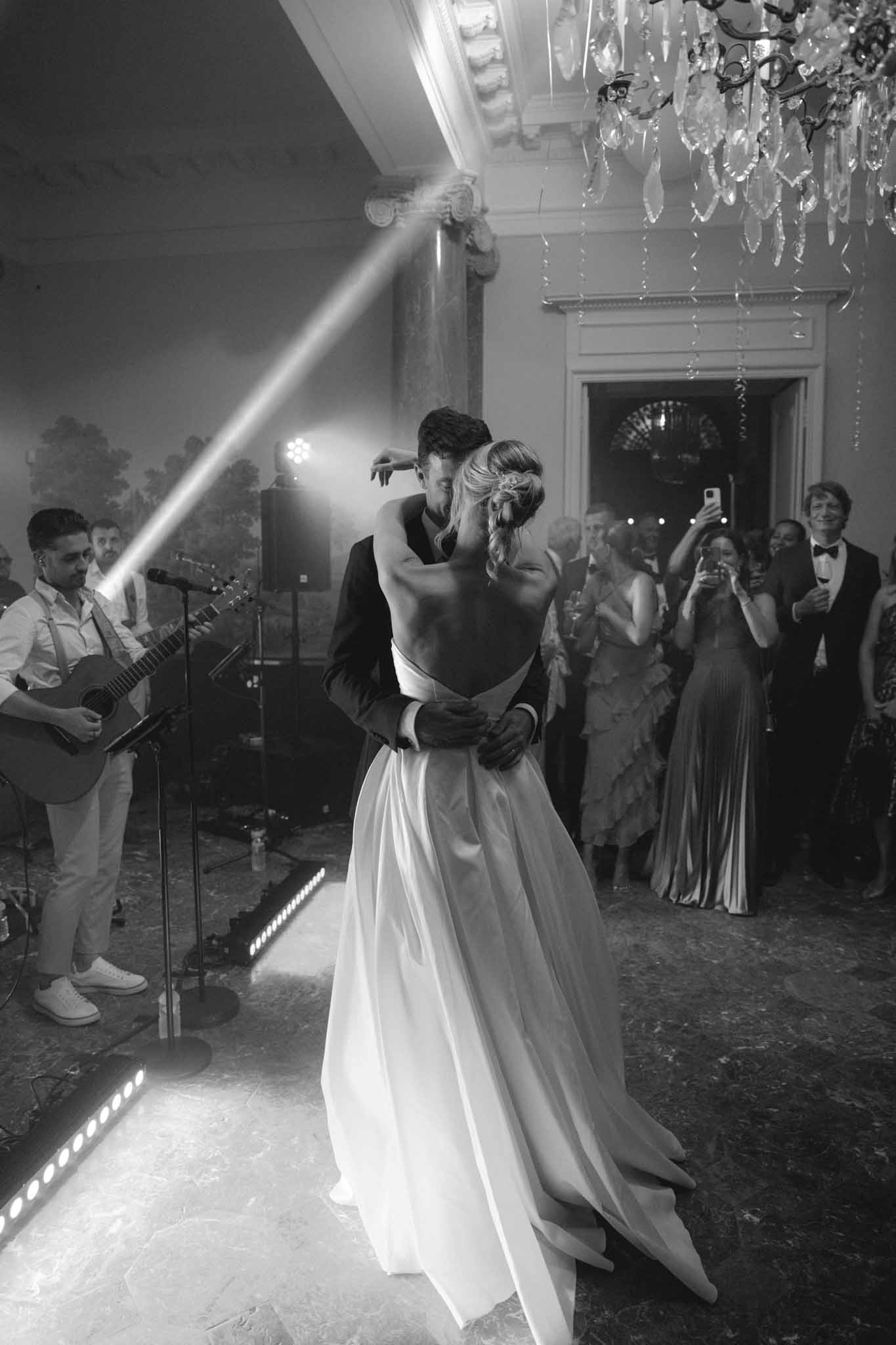 Bride and groom share their first dance under a crystal chandelier in a black-and-white ballroom photograph