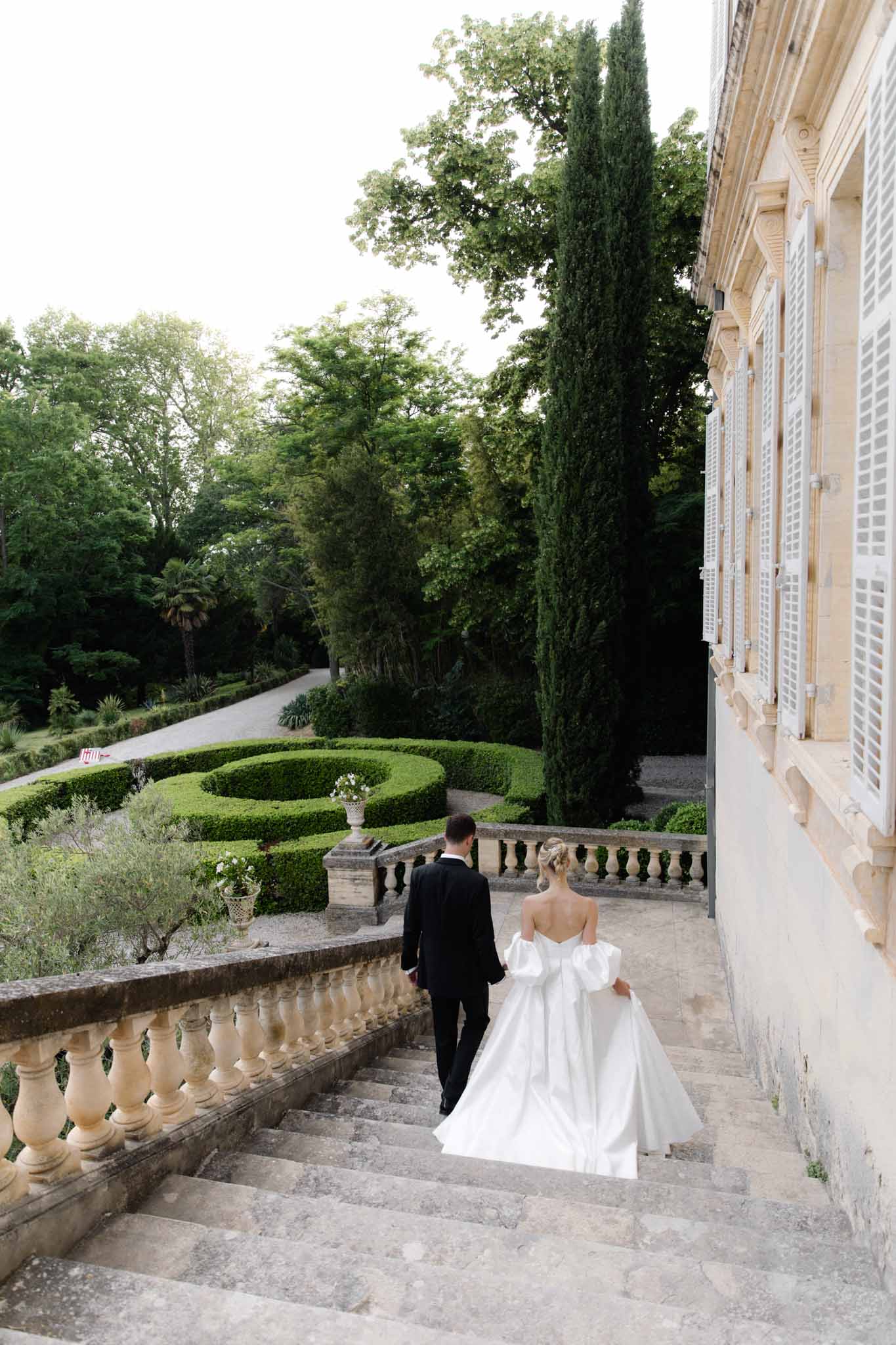 Bride and groom descending stone staircase toward formal gardens with cypress trees at classical pale stone estate