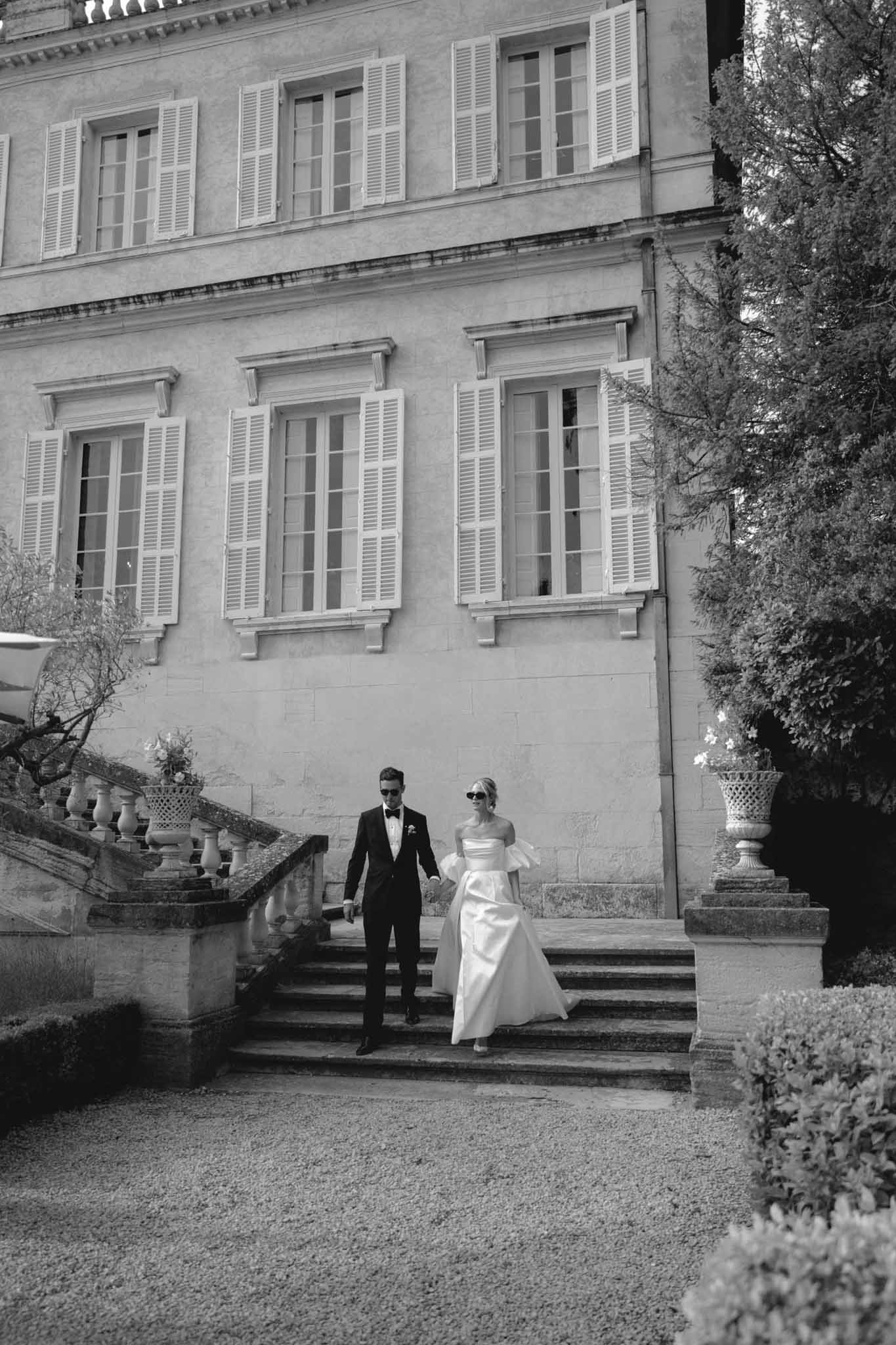 Black-and-white portrait of bride in off-shoulder gown and groom in tuxedo with sunglasses descending chÃ¢teau stone steps