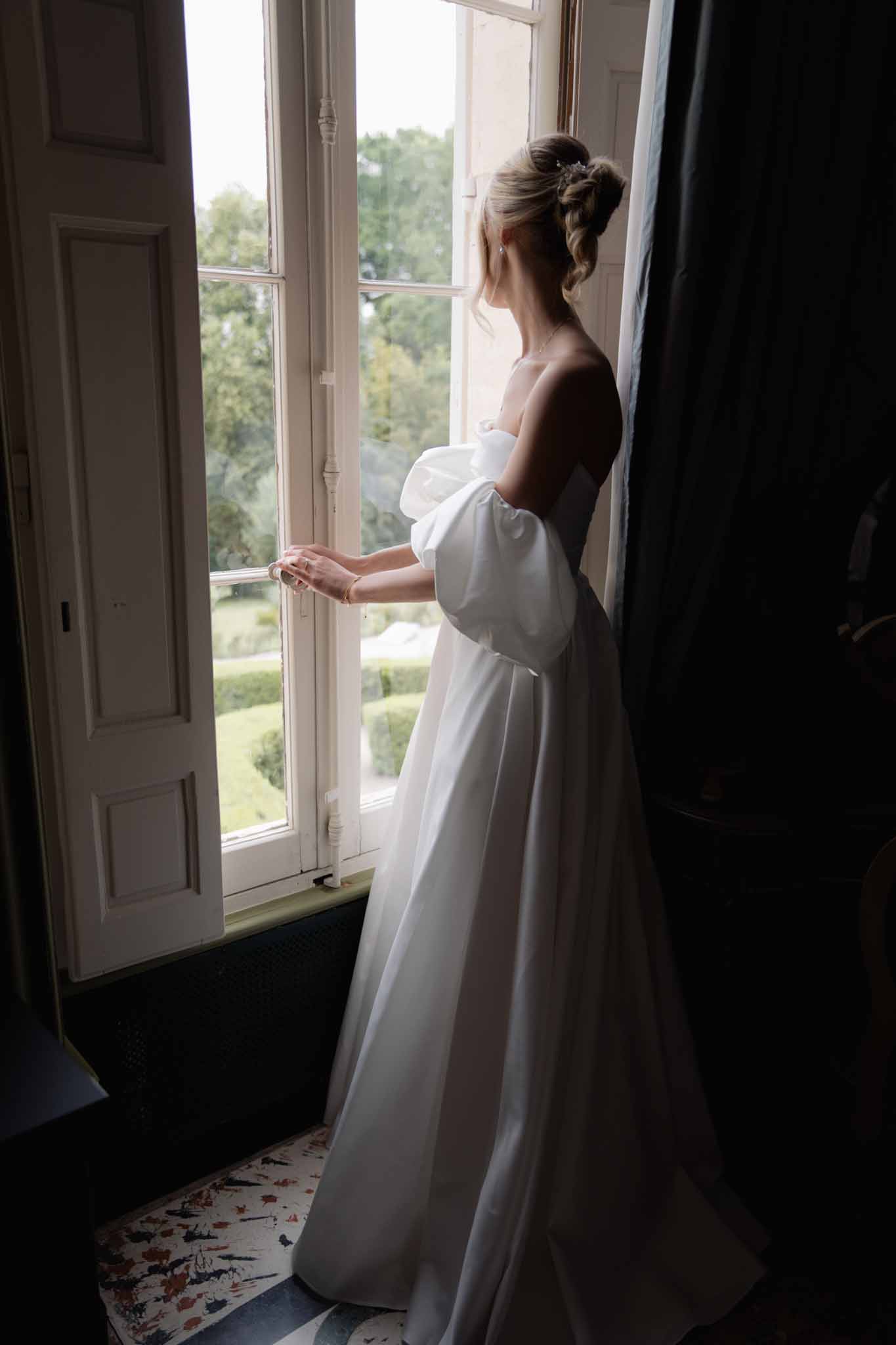 Bride in strapless ivory gown gazing out a tall window during getting-ready moment in a historic room