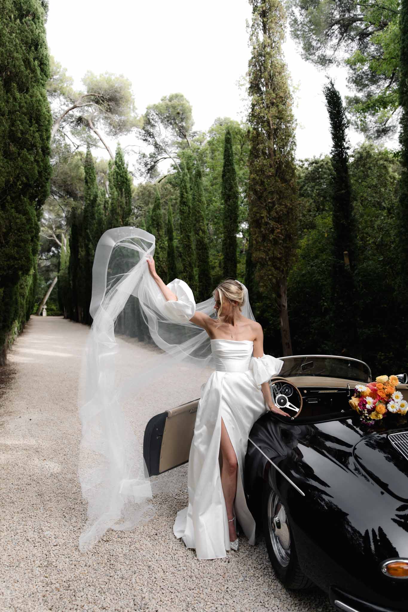 Bride in off-shoulder white silk gown beside a black convertible classic car on a cypress-lined gravel pathway, veil billowing in wind.