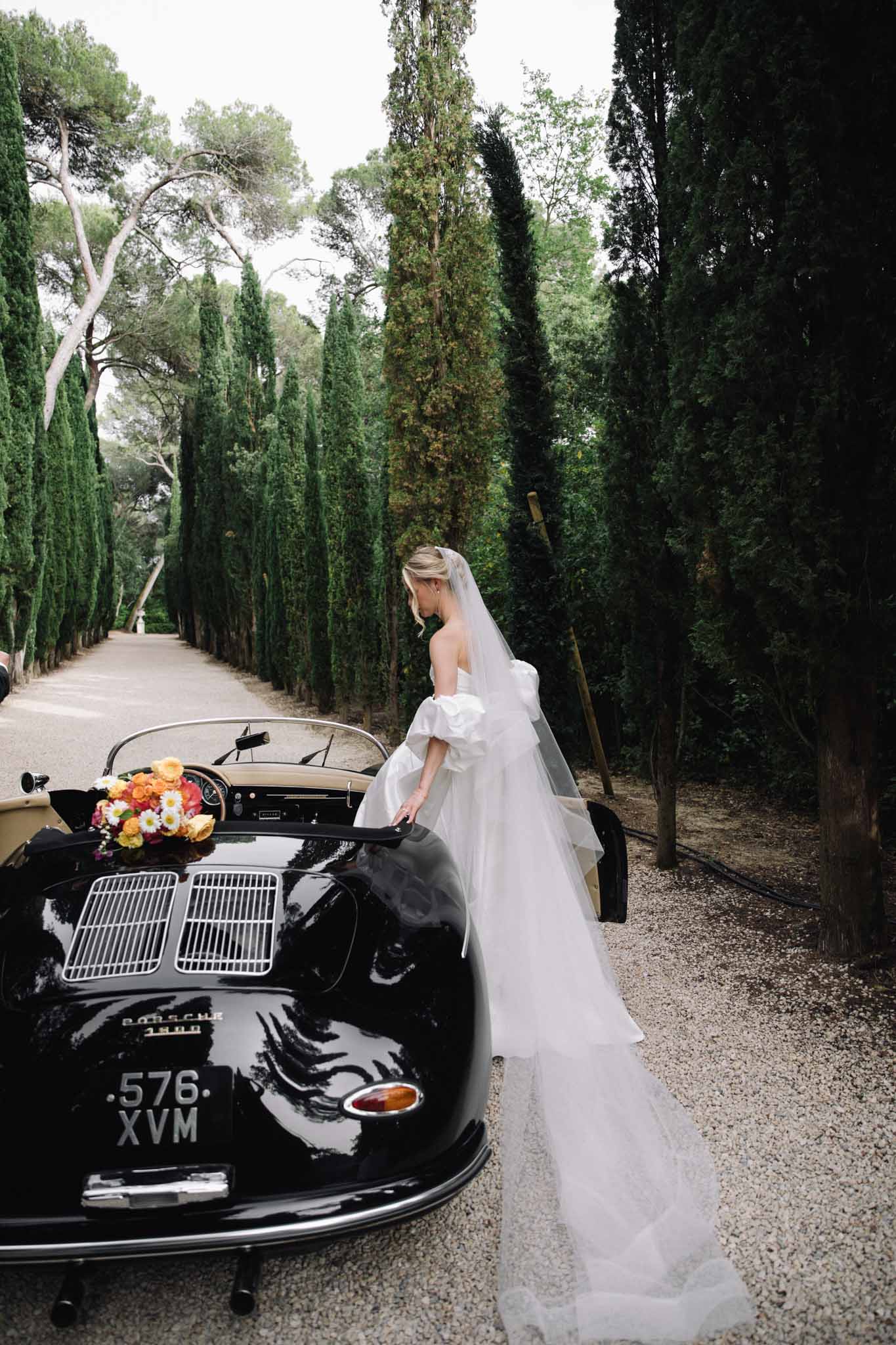 Bride in ivory off-shoulder gown with veil beside vintage black Porsche on cypress-lined driveway