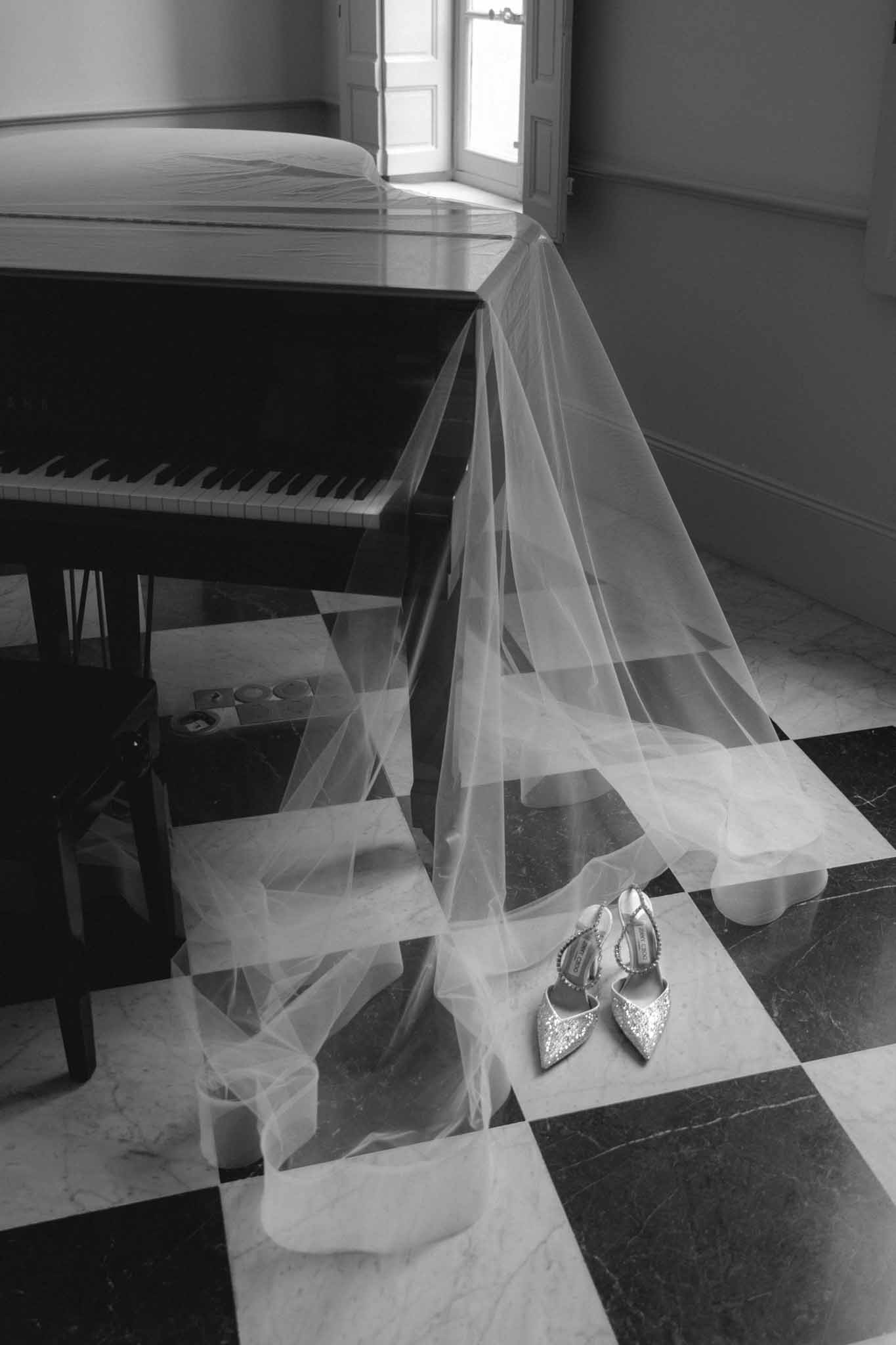 Black and white detail of bridal veil draped across floor with embellished shoes by grand piano