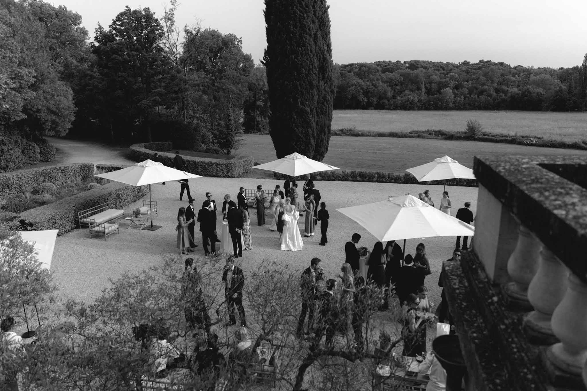 Aerial black and white view of cocktail hour on estate terrace with guests, white umbrellas, and cypress tree by water
