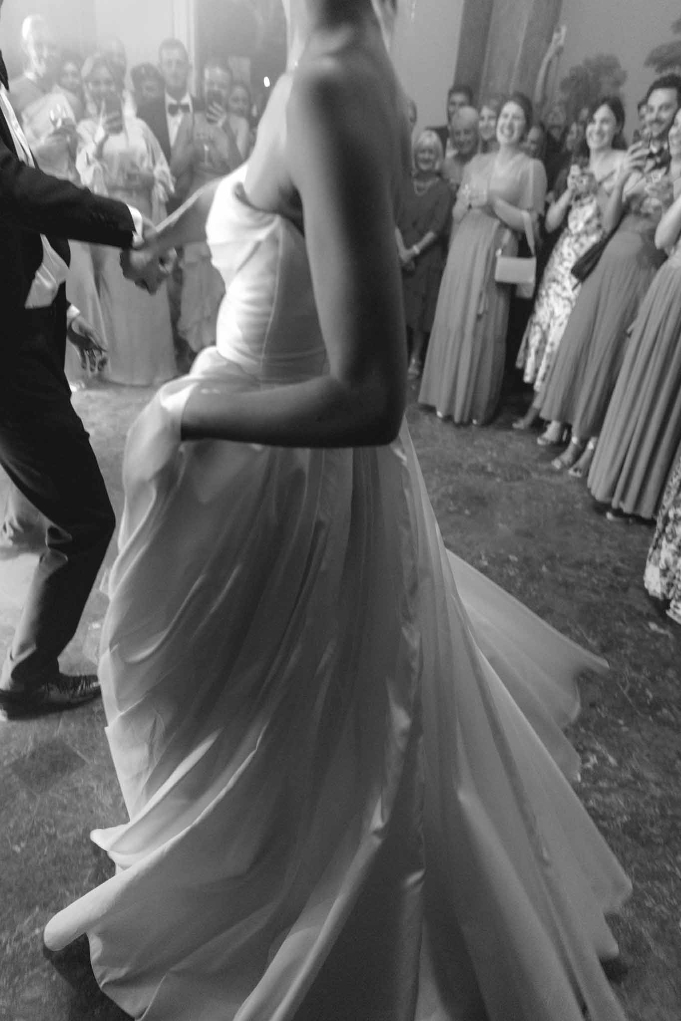 Black and white first dance with bride in strapless gown and groom in dark suit surrounded by watching guests