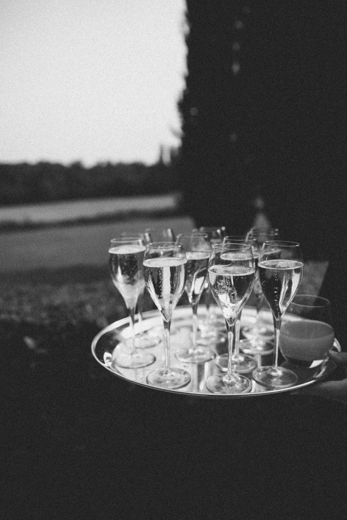 This black-and-white image shows a close-up detail shot of a round silver tray being carried by a server during what appears to be an outdoor cocktail hour. The tray holds approximately a dozen filled champagne flutes with visible bubbles, along with a small glass or cup to one side. The background is dark with high contrast, and a pale open landscape is visible in the upper left, suggesting the setting is an outdoor estate or château grounds at dusk or low light. The image has a grainy, film-like quality with deep shadow tones and bright highlights on the glassware.