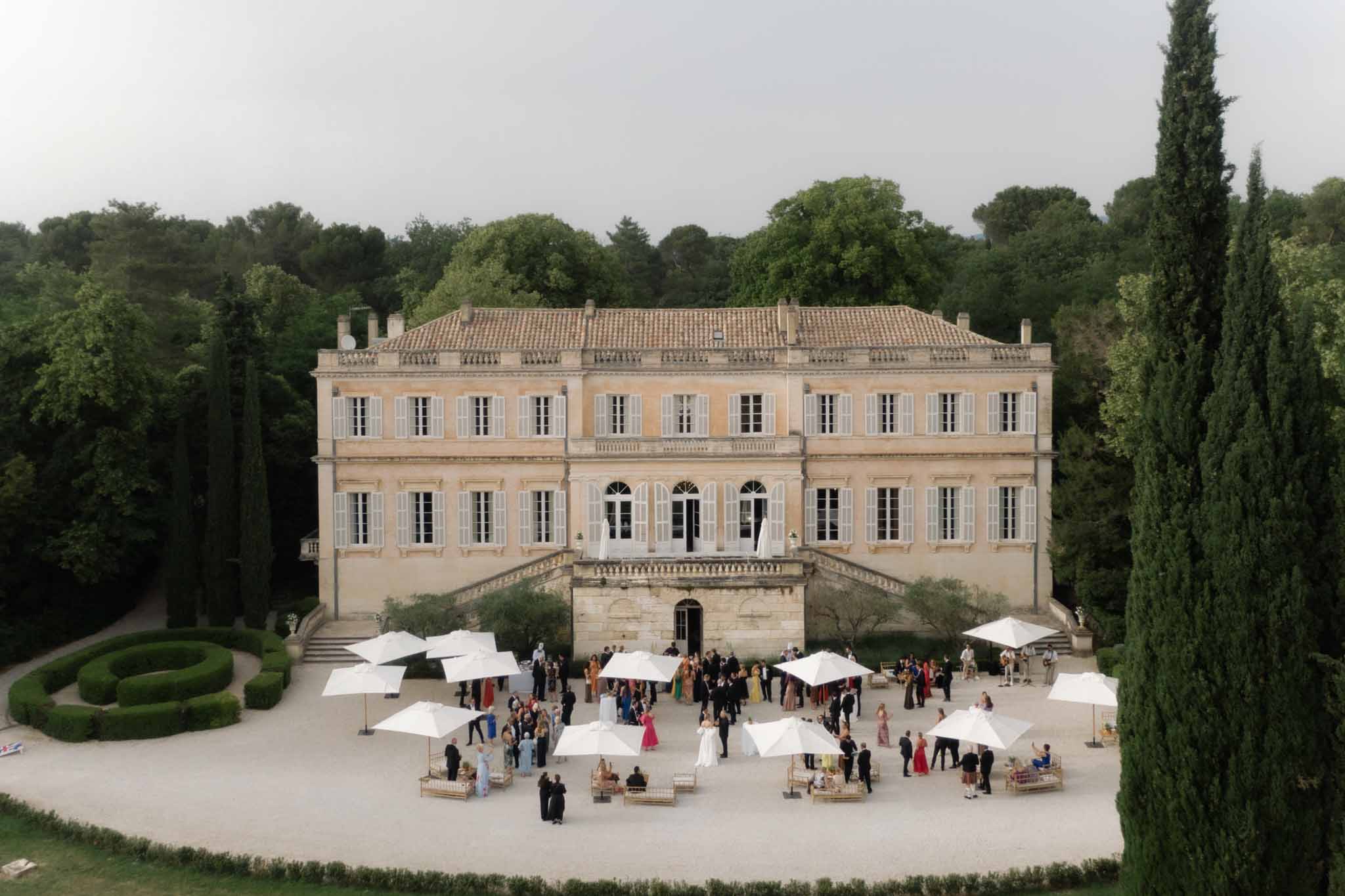 Aerial view of a classical French chÃ¢teau courtyard with 150 guests under white umbrellas, cypress trees flanking the facade.
