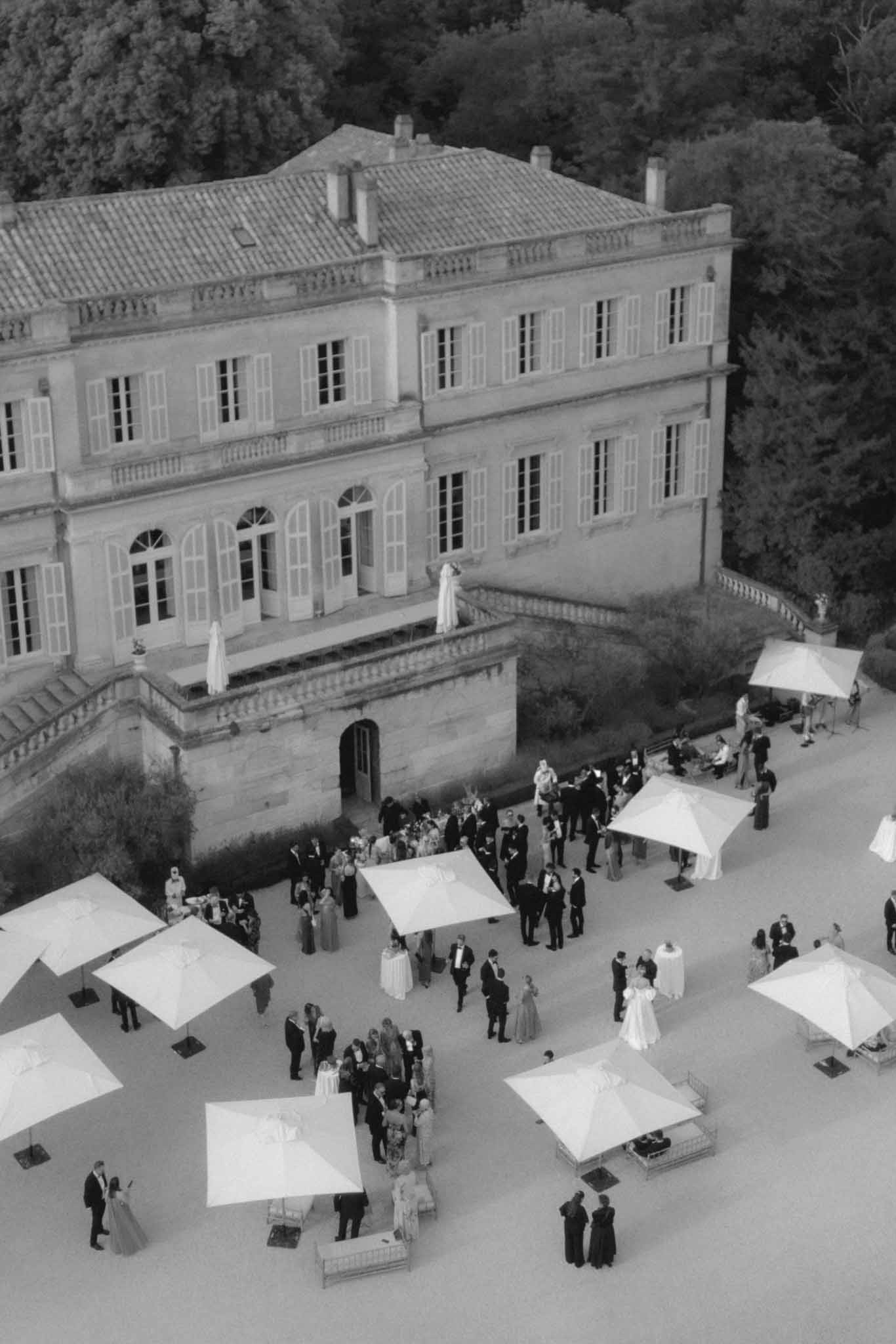 Aerial black-and-white view of wedding guests gathered in a classical chateau courtyard during cocktail hour
