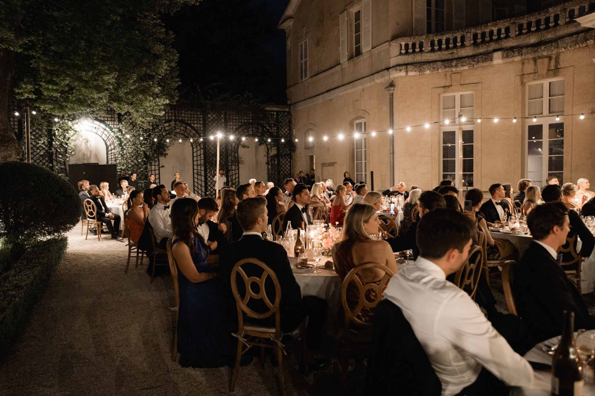Evening outdoor reception in chateau courtyard with round tables under string lights and guests watching a speech