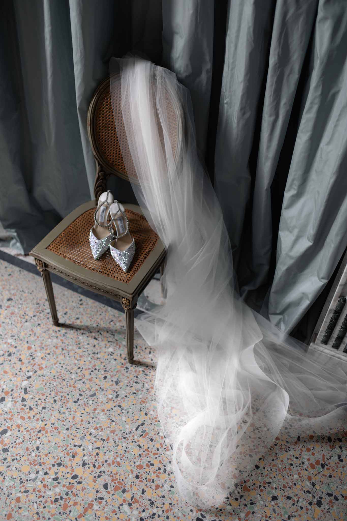 Bride's ivory tulle veil and crystal-embellished heels on a vintage rattan chair with sage green curtain backdrop