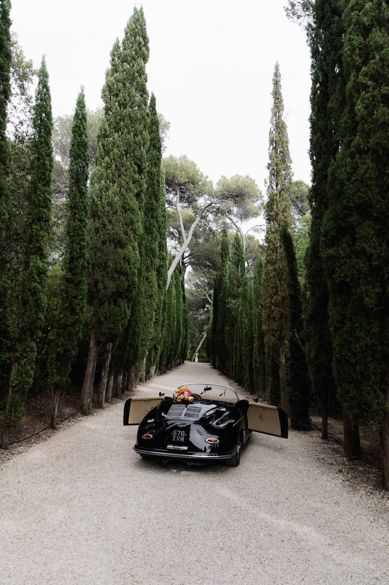 A vintage black convertible sports car — resembling a classic Porsche Speedster, with a French license plate reading 576 XVM — is parked on a gravel driveway with both doors open, viewed from the rear in a wide shot. A colorful bridal bouquet featuring orange, red, and yellow blooms is visible resting on the car. The driveway is lined symmetrically with tall Italian cypress trees forming a dramatic corridor that recedes into the distance, characteristic of a formal French estate or domaine approach. The overall styling leans classic and understated, with the black-and-cream car interior serving as the main intentional design element in the frame. Potential venue feature image.