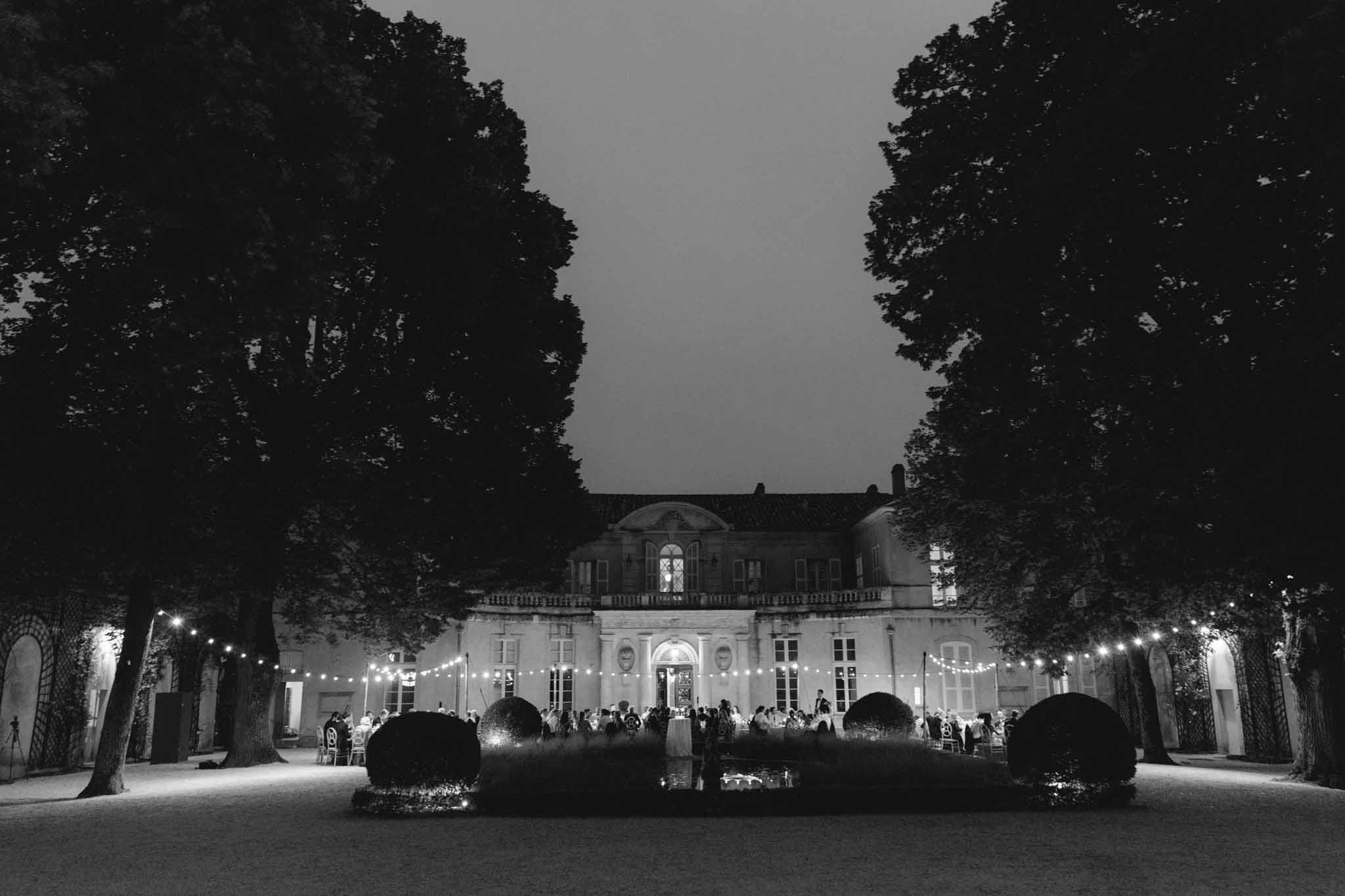 Outdoor night reception with guests at tables before uplift chateau facade with bistro lights in B&W