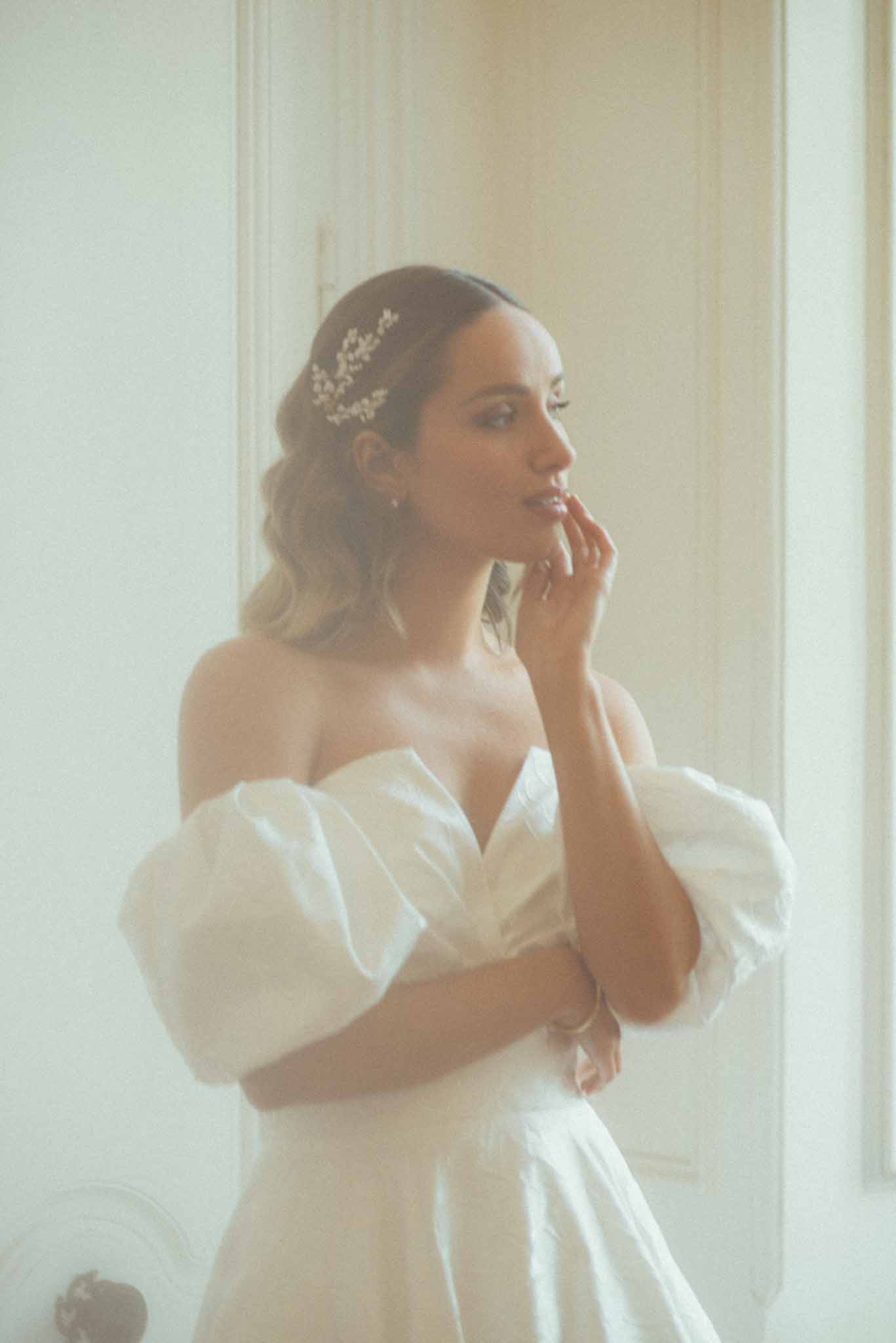 A bridal portrait shot indoors, likely in a château or period building suggested by the white panelled door and ornate moulding visible in the background. The bride stands in three-quarter profile, one hand raised to her lips, wearing a white off-the-shoulder gown with voluminous puff sleeves and a sweetheart neckline. Her hair is worn half-up with loose waves, accessorised with a delicate gold and pearl floral hair comb. The image is softly overexposed with a bright, airy quality, and the composition is a close-up portrait focused from the waist up.