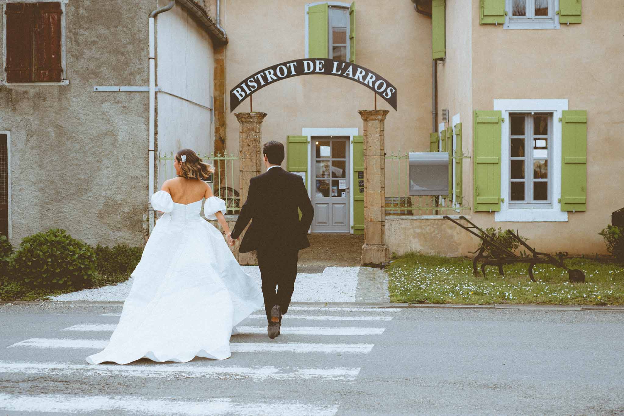 A couple portrait shot taken outdoors on a village street, showing the bride and groom walking hand-in-hand across a pedestrian crossing toward the entrance of a building signed 'Bistrot de l'Arros.' The bride wears a white off-the-shoulder ball gown with voluminous puff sleeves and a full skirt with a short train, her hair worn down with a small white floral hair accessory. The groom is dressed in a dark navy or black suit. The building facade is pale yellow with sage green shutters and a stone arched entrance gate bearing the bistrot's name. The image is a wide shot taken from behind the couple, capturing the full length of the dress and the village setting. The overall style is classic French countryside.
