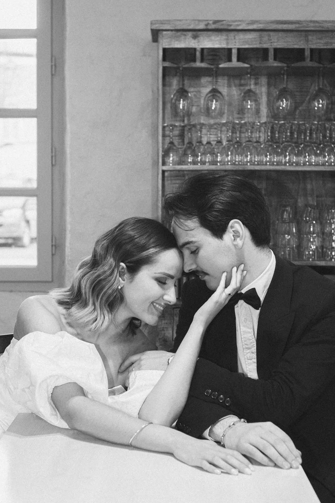 This is a black-and-white couple portrait shot indoors, likely at a bar or cellar area of a venue. The bride and groom are leaning close together with foreheads nearly touching, the bride resting her arms on a white surface while gently placing her hand on the groom's face. The bride wears an off-the-shoulder dress with puffed sleeves and has short, wavy hair accessorized with small stud earrings; she is smiling softly with eyes closed. The groom wears a dark suit jacket with a bow tie and white dress shirt, and has a thin mustache. In the background, a rustic wooden wine glass rack holds rows of stemware hung upside down, adding a distinctly French venue character to the setting. The image has soft, bright tones with gentle contrast, giving it a film-like quality. The composition is a close portrait framing both subjects from roughly the waist up.