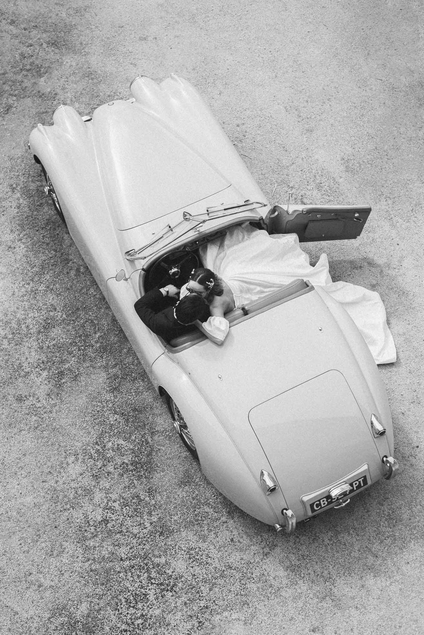 Overhead black-and-white shot of bride and groom kissing inside a vintage 1950s open-top convertible car