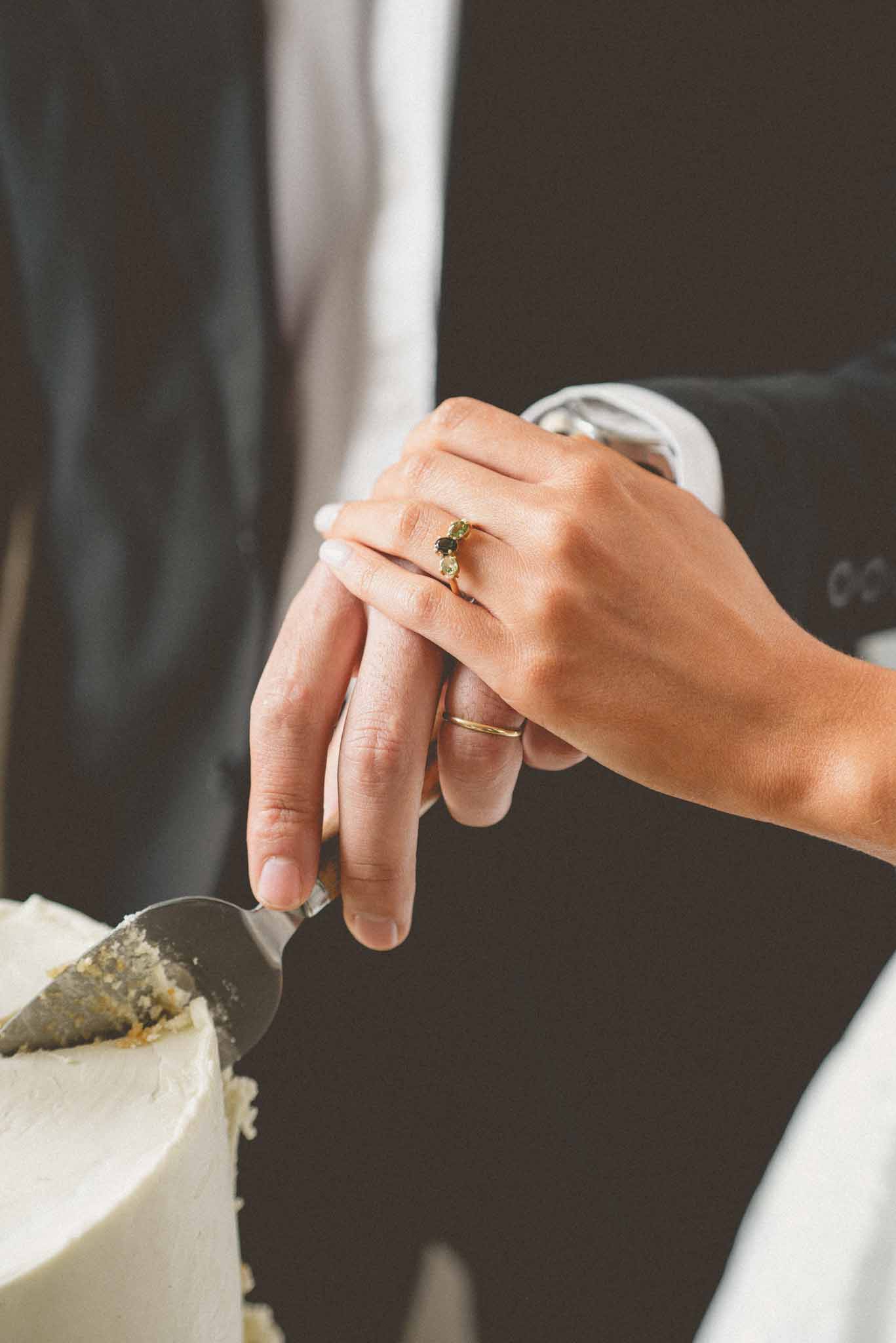 Close-up detail shot of a couple cutting their wedding cake together, with both hands placed on a silver cake server. The cake is white with smooth frosting, partially cut to reveal a golden sponge interior. The bride wears a gold engagement ring set with a dark center stone flanked by two pale green stones, along with a slim gold wedding band. The groom wears a plain gold wedding band and a silver watch, and is dressed in a dark charcoal suit with a white dress shirt visible at the collar. The background is dark and out of focus, keeping all attention on the hands and rings.