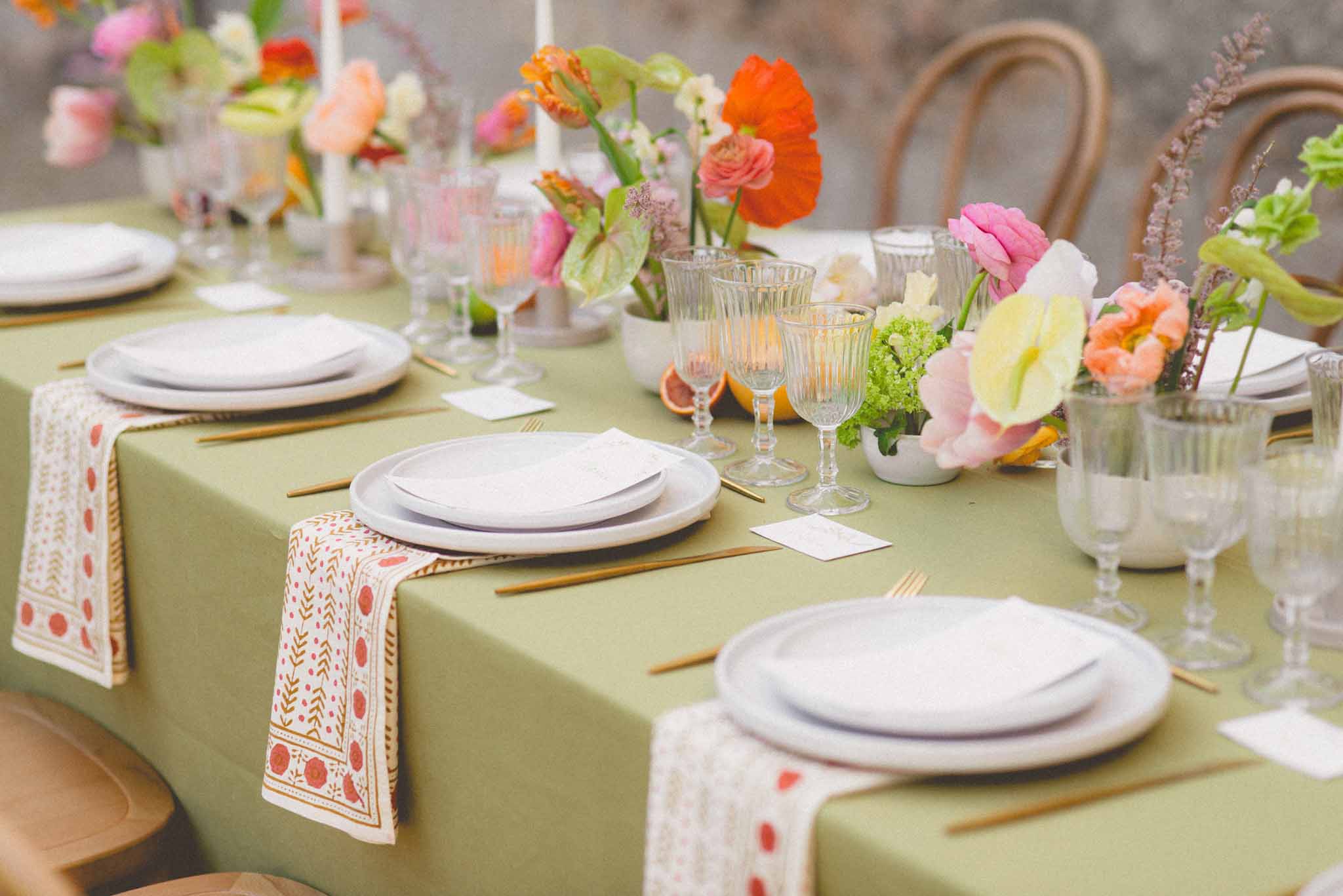 Reception table with sage green linen, block-print napkins, orange poppies, pink ranunculus, and citrus accents