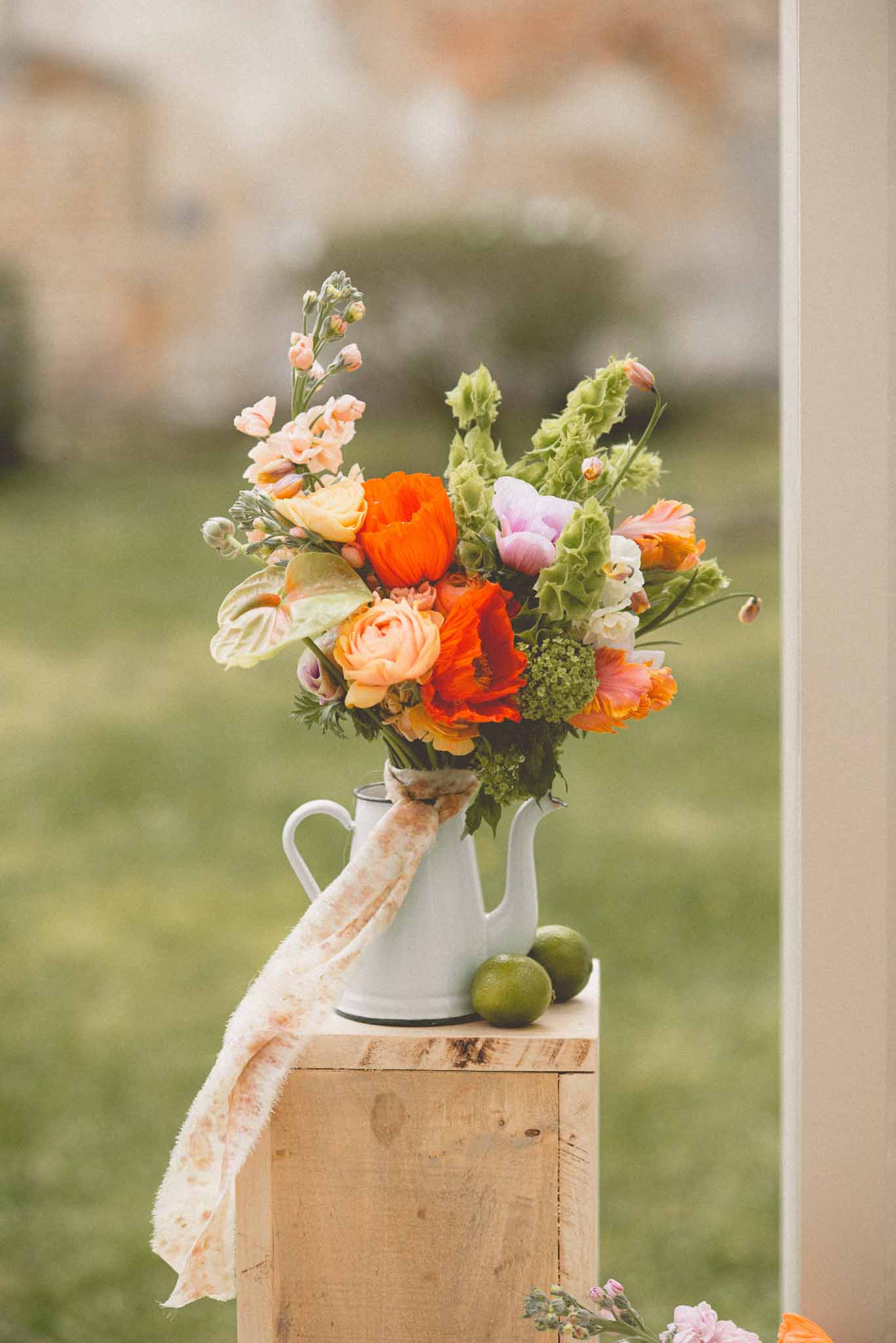 Bohemian floral arrangement with orange poppies and peach roses in white enamel coffee pot on wood crate