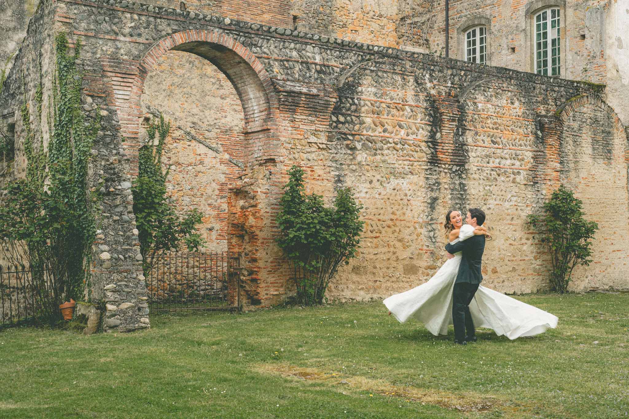 Groom lifting and spinning bride in ivory ball gown against ancient stone wall of a French chateau