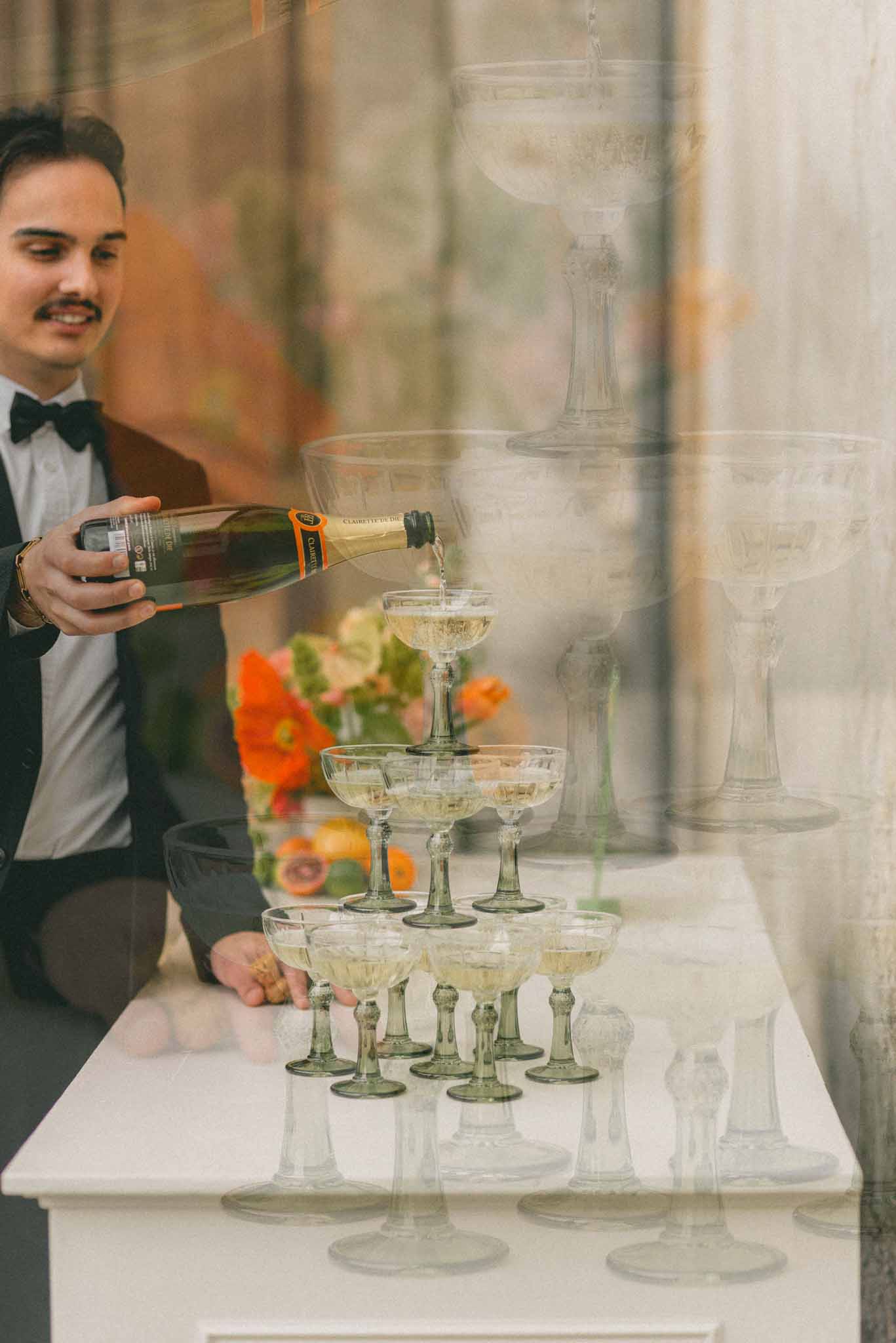 Man in black tuxedo pours champagne into smoke-green coupe glass tower with orange florals in background