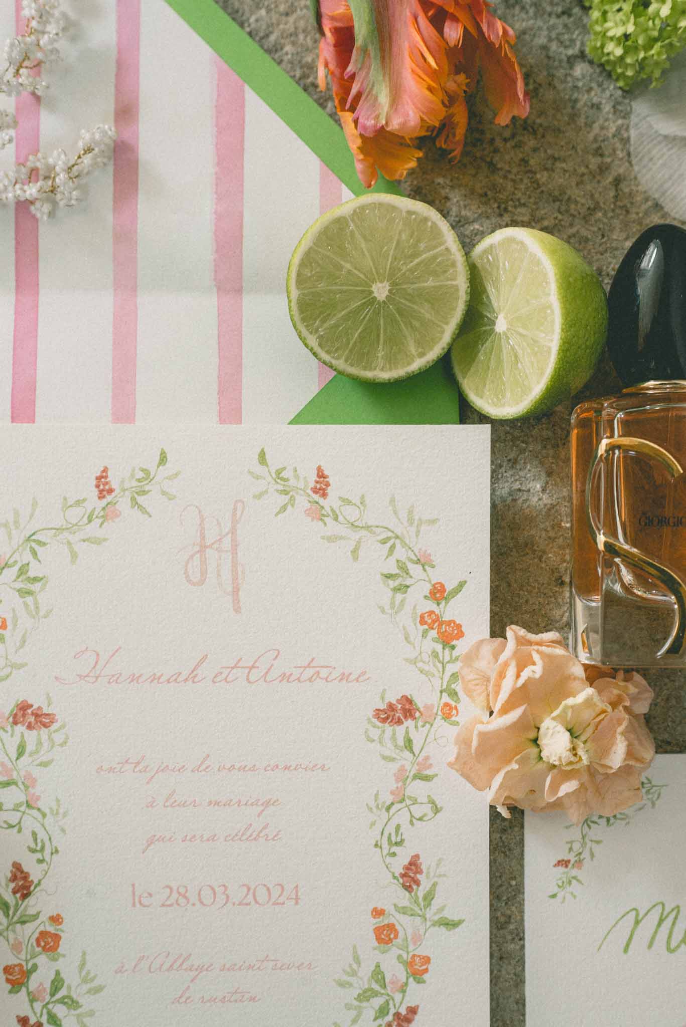 A close-up flat lay detail shot of a wedding stationery suite for Hannah et Antoine, dated 28.03.2024, with the venue listed as l'Abbaye Saint-Sever de Rustan. The invitation card features hand-painted watercolor floral border illustrations in coral, terracotta, and sage green with a calligraphy script in dusty pink and green tones. Surrounding the stationery are styled props including two halved limes on a bright green envelope, an orange parrot tulip, a peach garden rose bloom, small white flowering branches, and a Giorgio perfume bottle. A secondary card with pink and white vertical stripes is partially visible in the upper left corner. The overall color palette of the flat lay is coral, peach, lime green, and pink, suggesting a vibrant, garden-inspired wedding aesthetic.