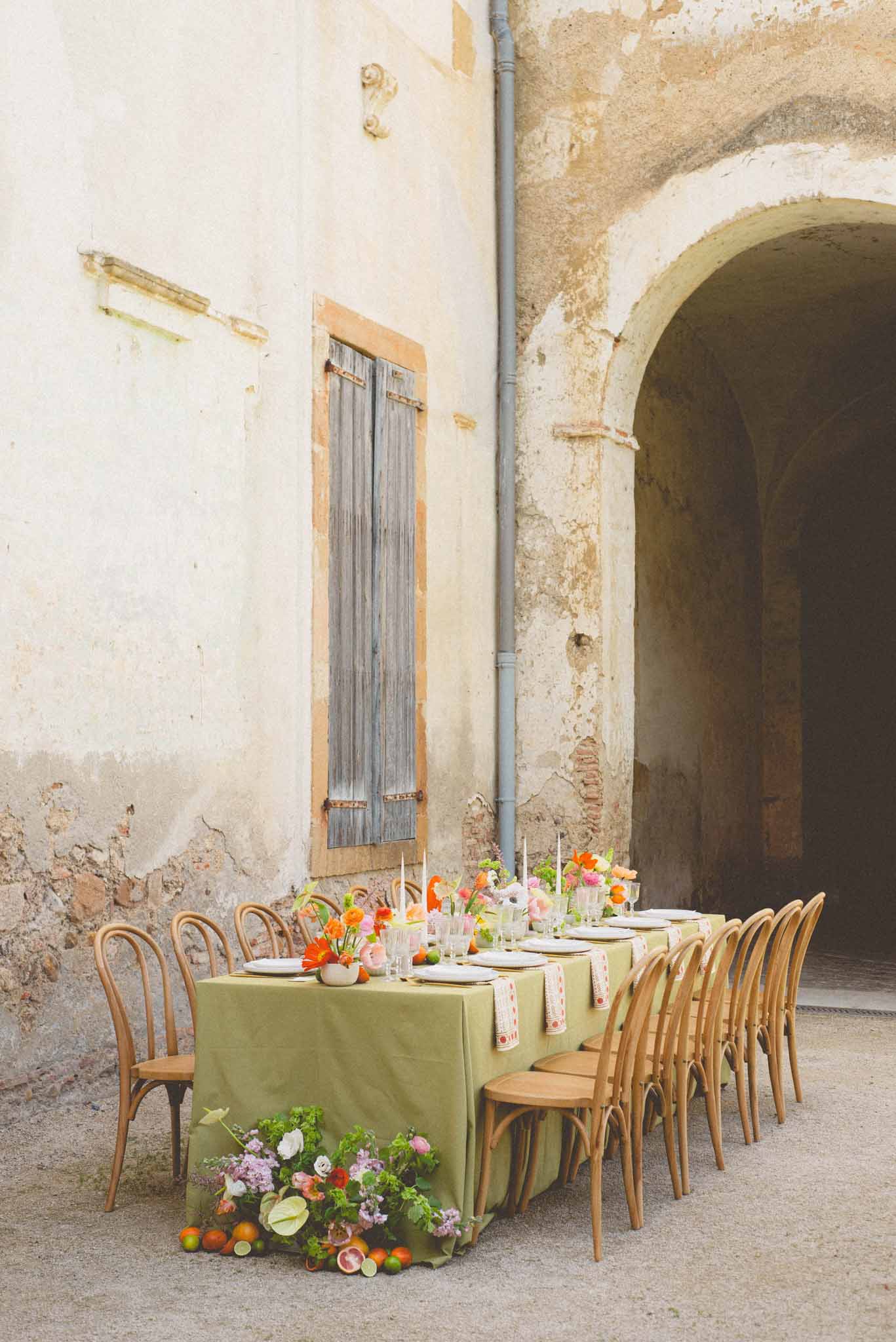 Olive linen table with orange poppies, pink ranunculus, and citrus cascading before weathered stone facade