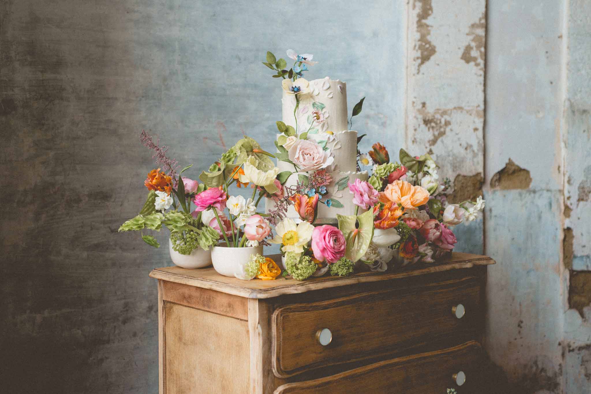 Three-tier painted buttercream cake surrounded by pink ranunculus, orange tulips, and yellow daffodils