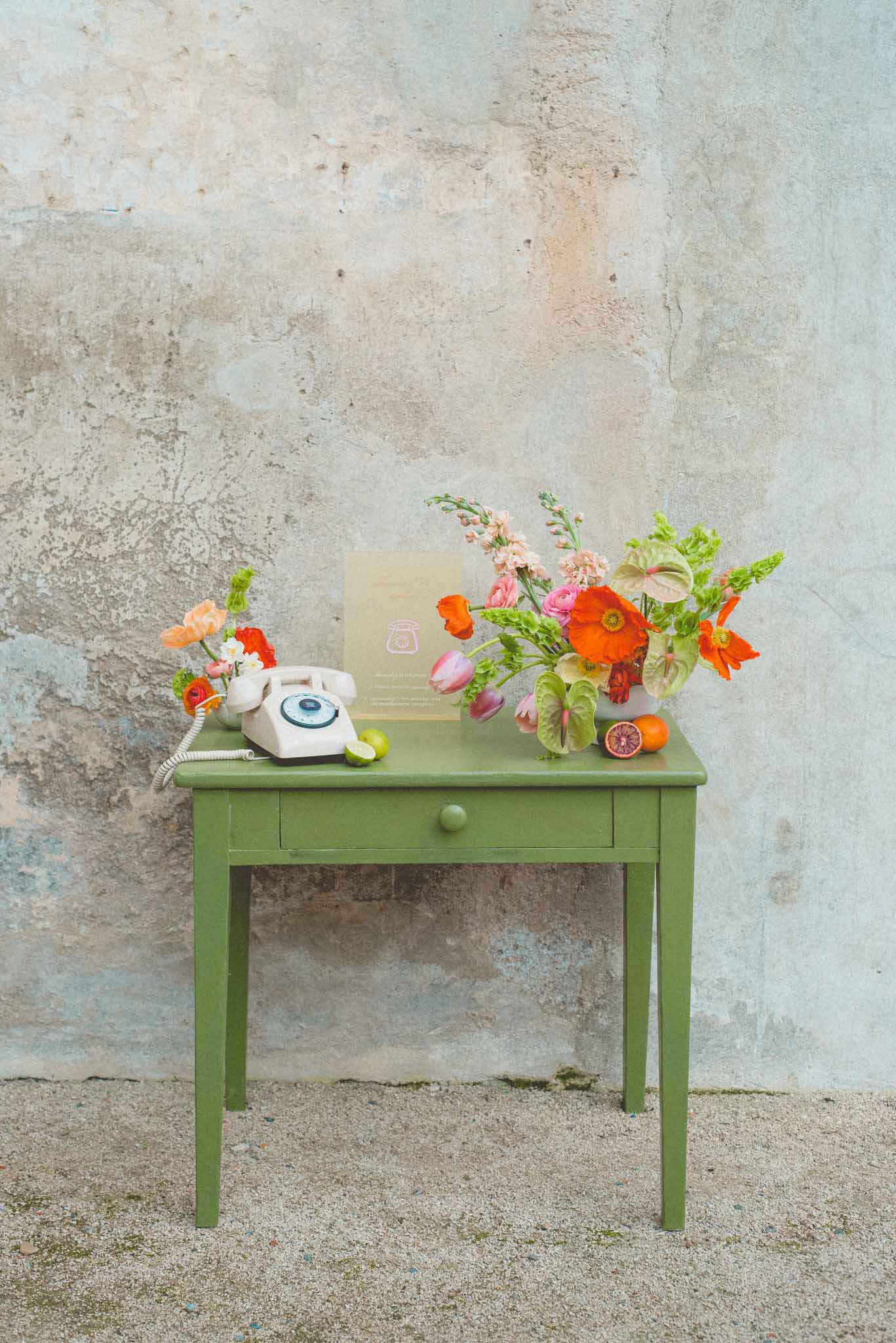 Styled vintage telephone guest book station with orange poppies, pink tulips, and citrus fruit on green side table