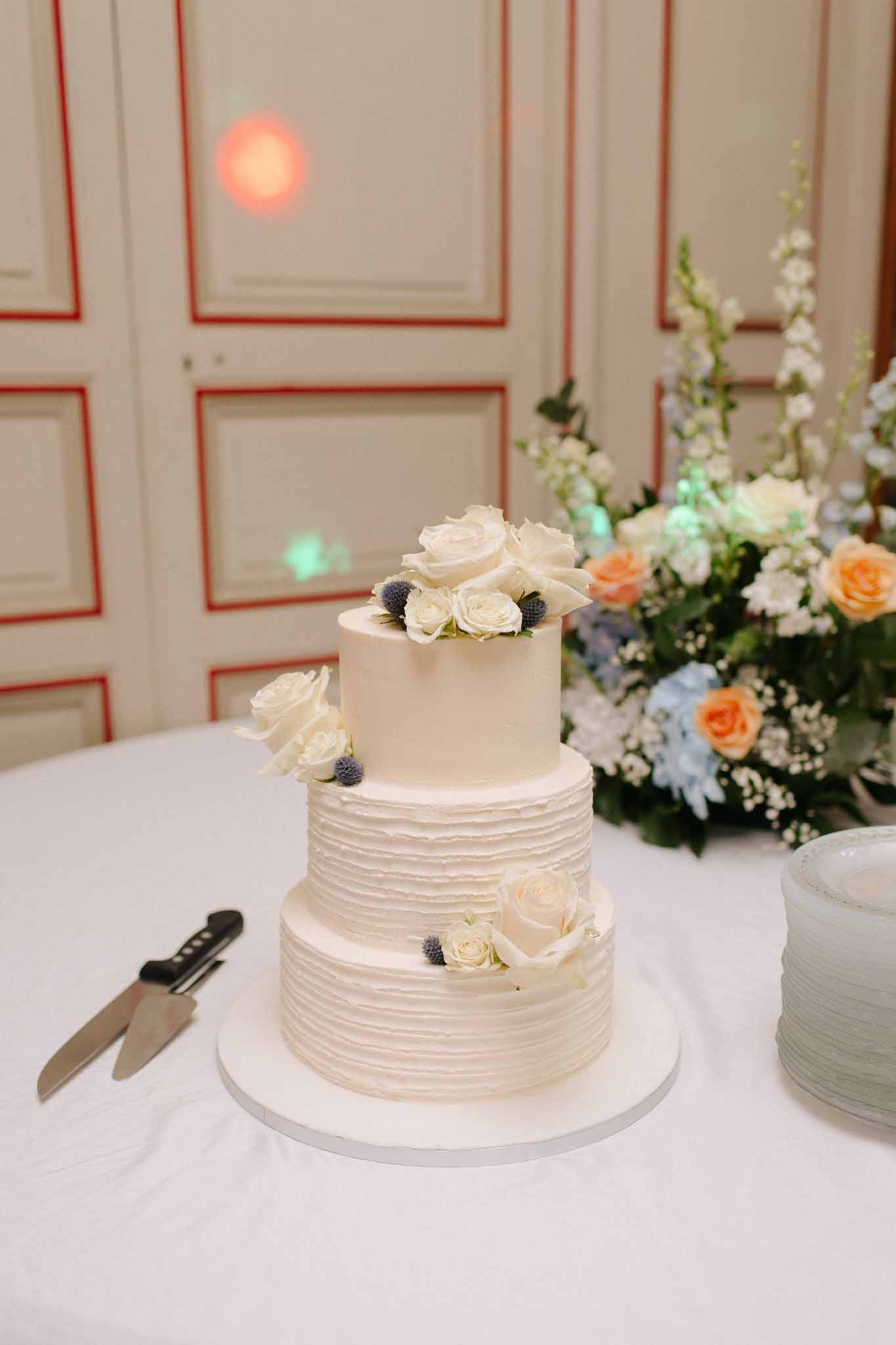Three-tiered ivory wedding cake with garden roses beside tall arrangement of peach roses and blue delphinium