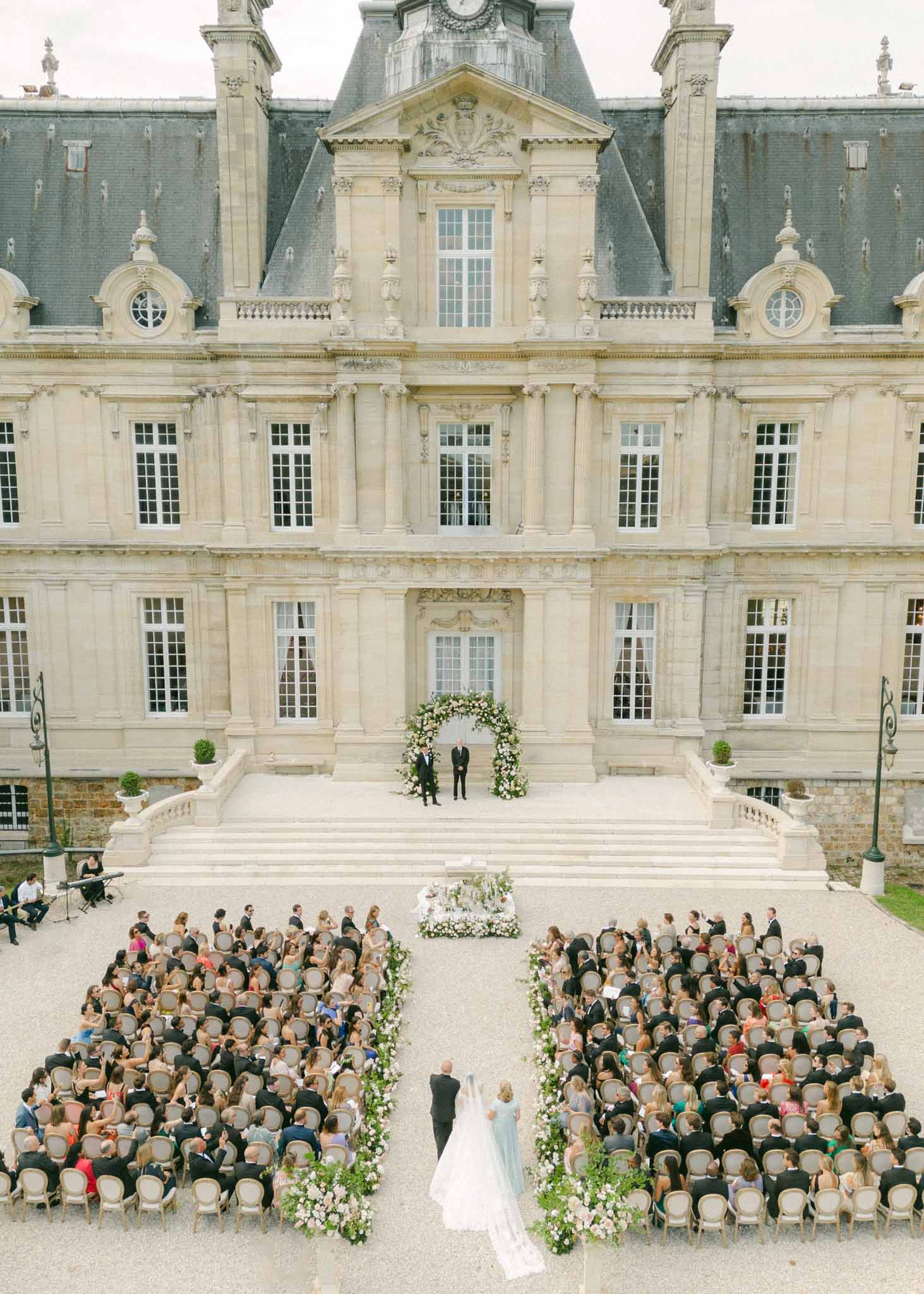 Aerial view of bride walking aisle toward groom under floral arch, 150 guests seated in courtyard of cream stone chÃ¢teau
