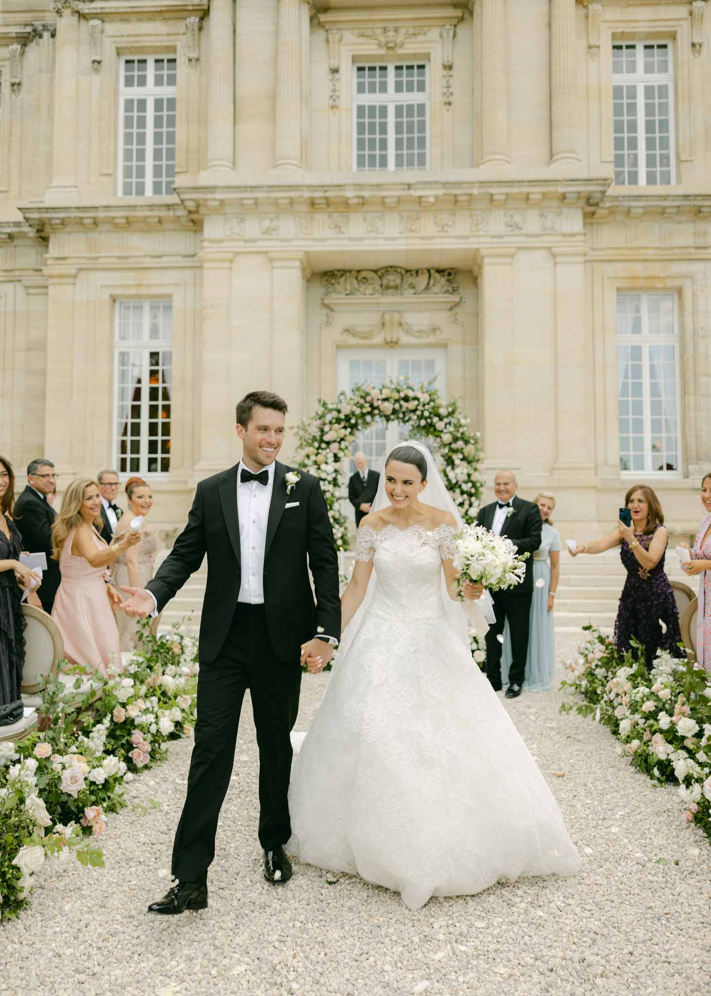 Bride and groom walking the recessional through a stone courtyard, guests applauding on both sides, white floral arch behind.