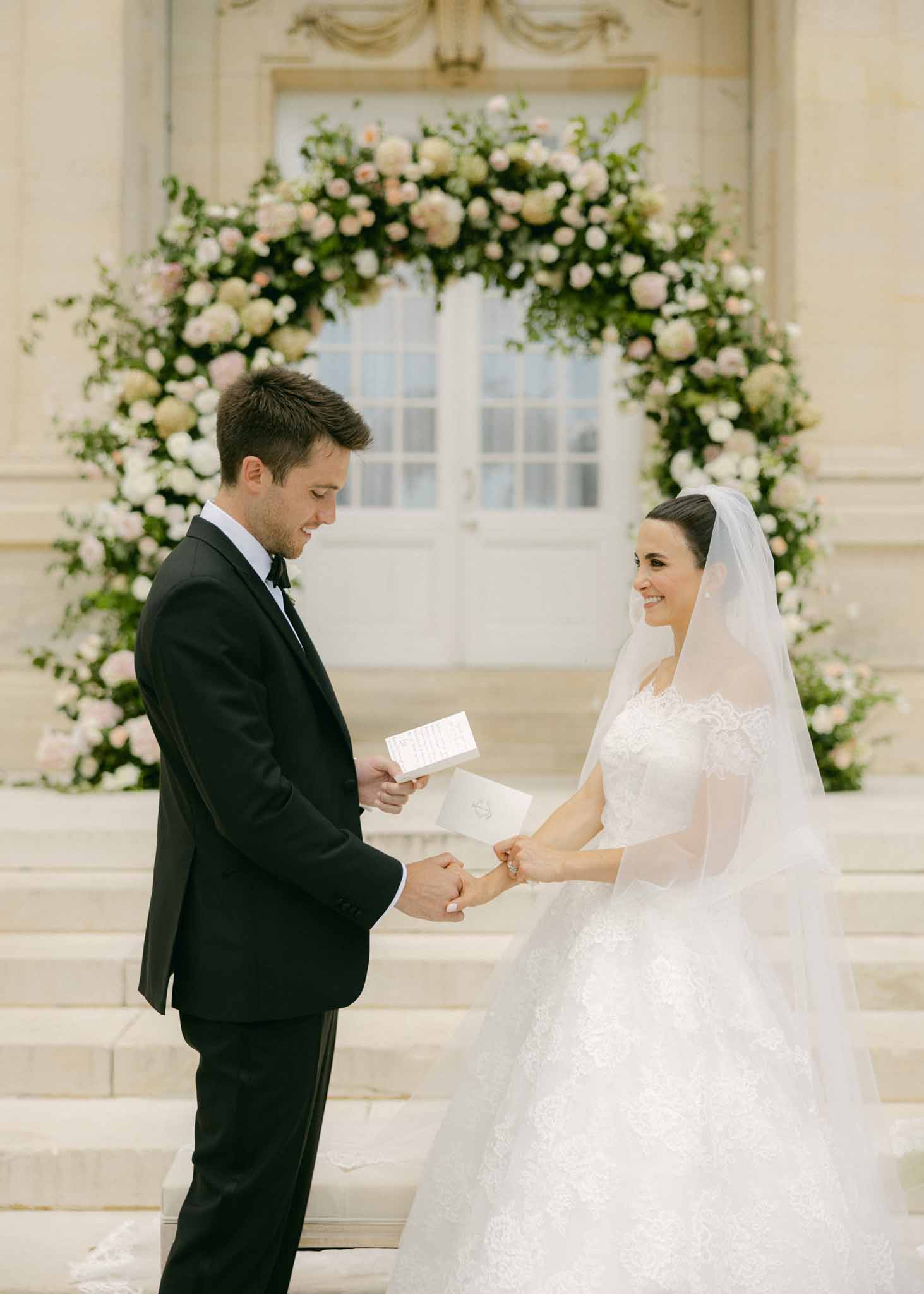 Bride and groom exchange rings under floral arch of cream and pink roses in elegant indoor ceremony with French doors