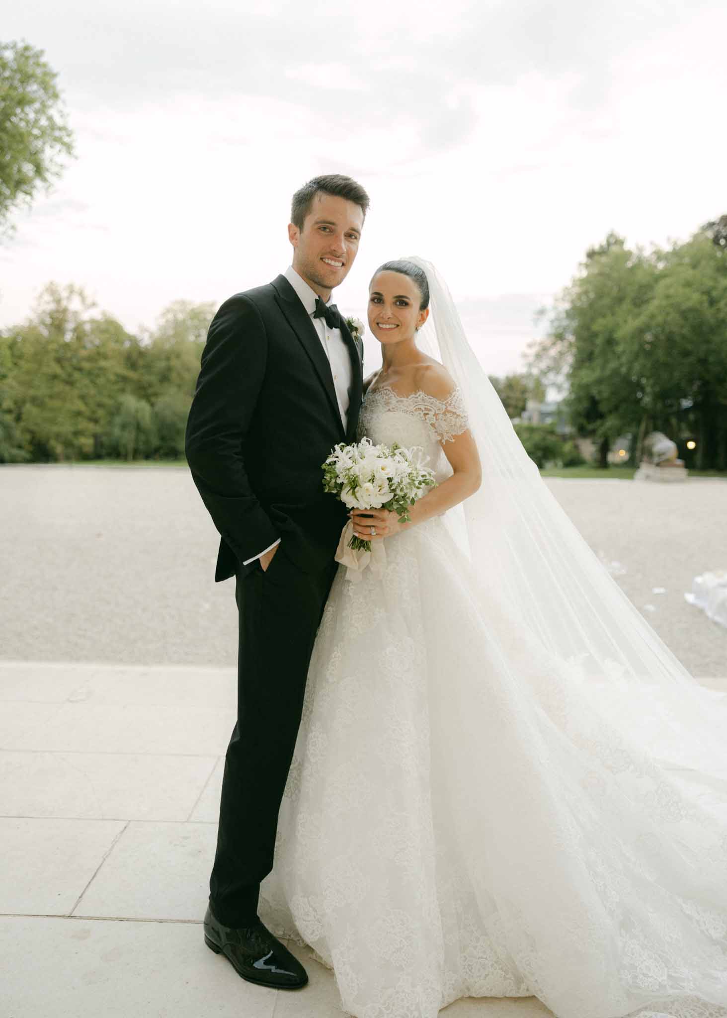 Formal portrait of bride in ivory lace gown with long veil and groom in tuxedo on garden terrace