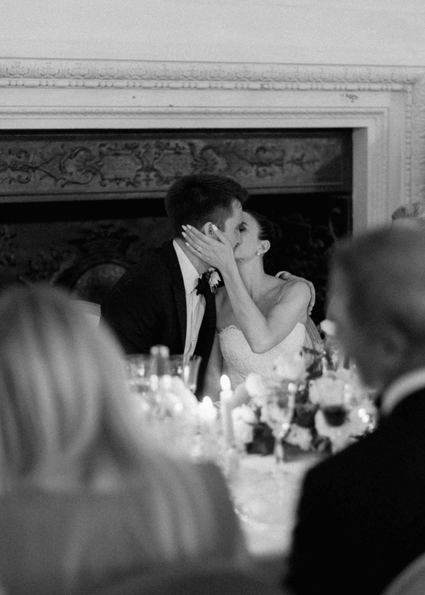 Black and white photo of bride and groom kissing at reception table beside ornate fireplace mantel