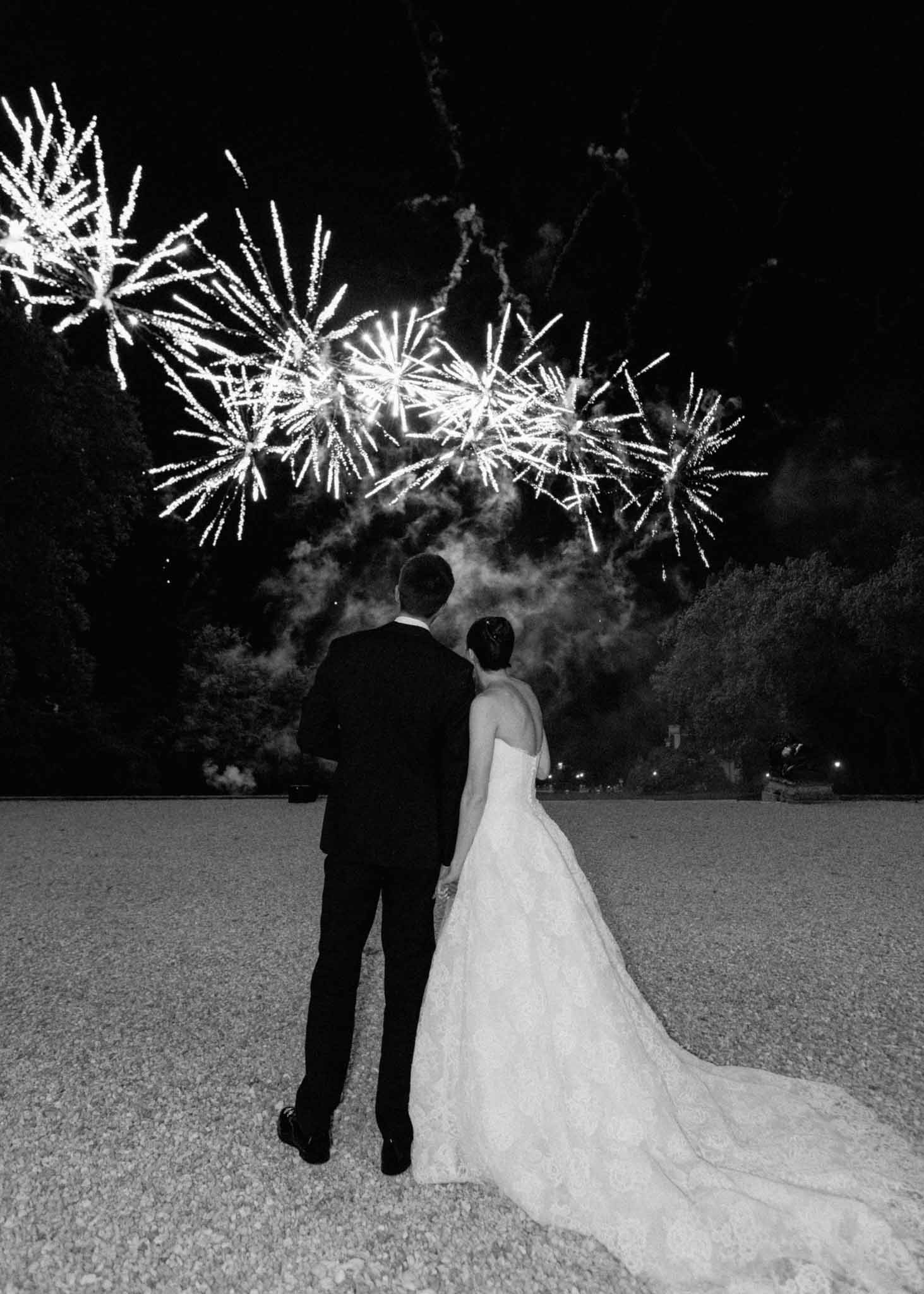 Black and white photo of bride and groom watching fireworks on outdoor terrace at night