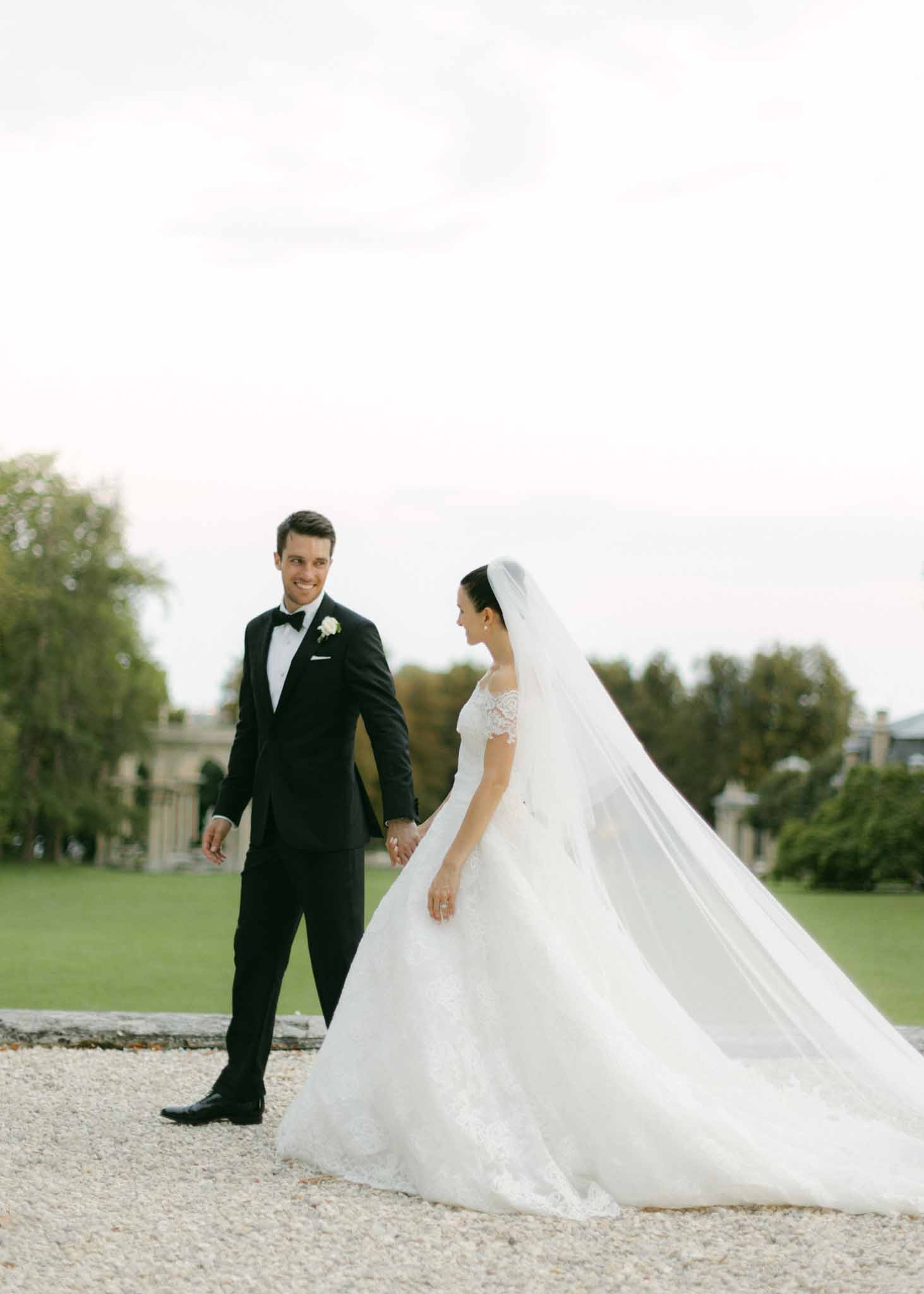 Bride in ivory lace ball gown with long veil and groom in black tuxedo hold hands on gravel path at estate
