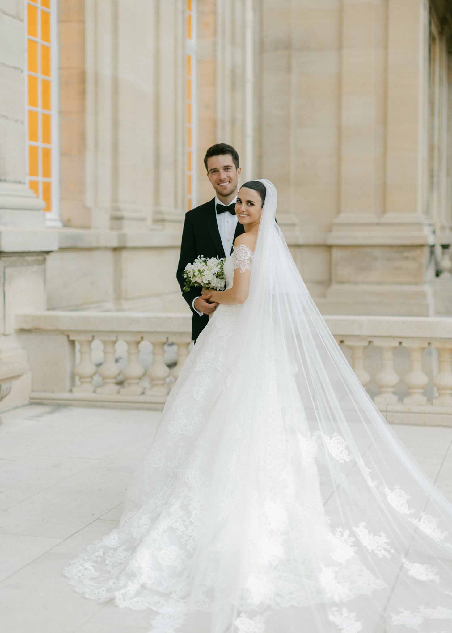 Bride in ivory lace gown with cathedral veil and groom in tuxedo posed before neoclassical columns