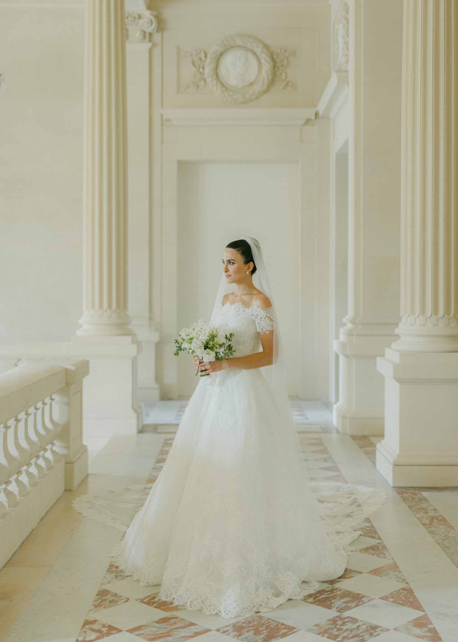 Bride in ivory lace off-shoulder gown holding white bouquet, standing in neoclassical corridor with fluted columns
