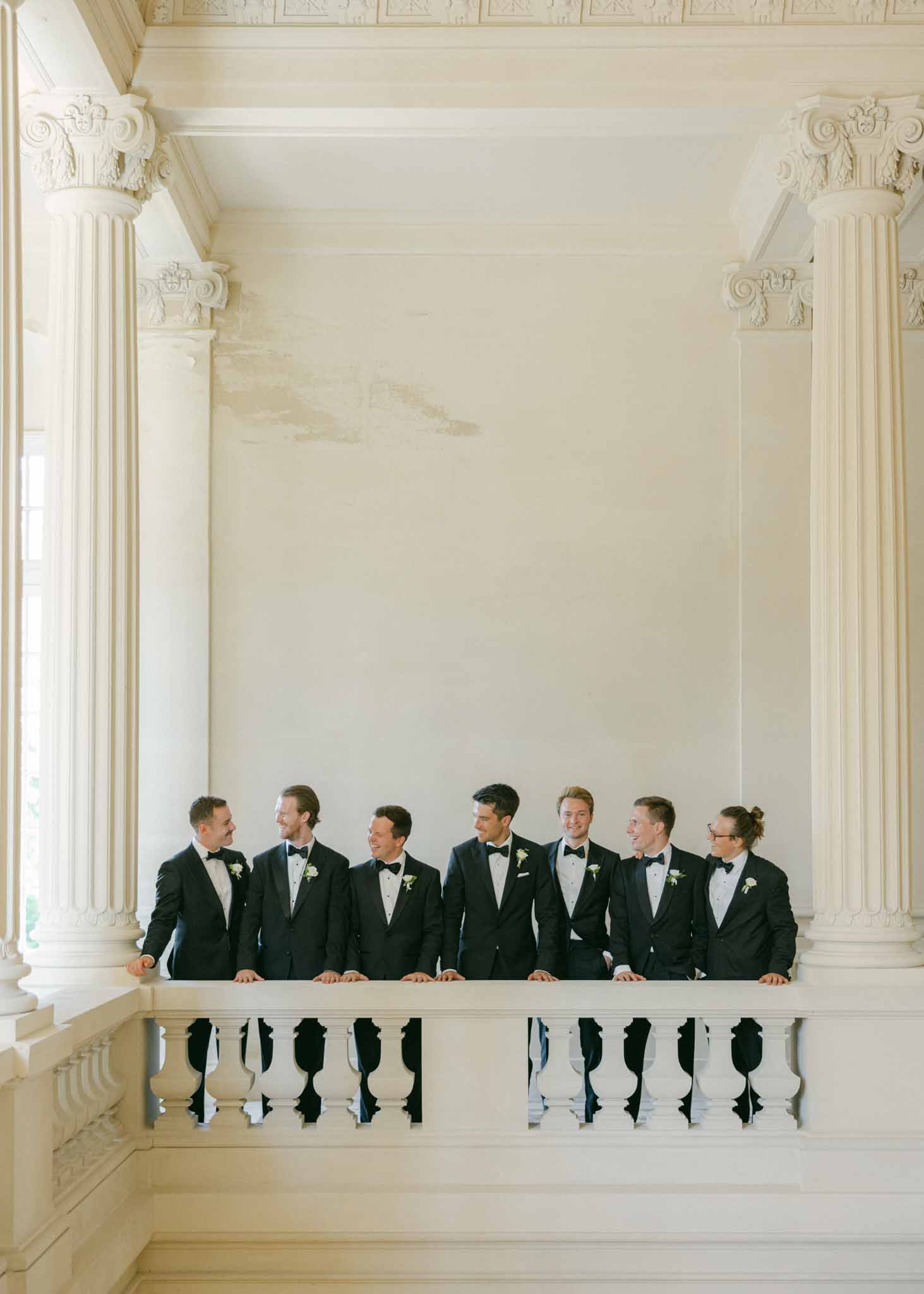 Seven groomsmen in black tuxedos on classical interior balcony between tall Corinthian columns with ornate capitals