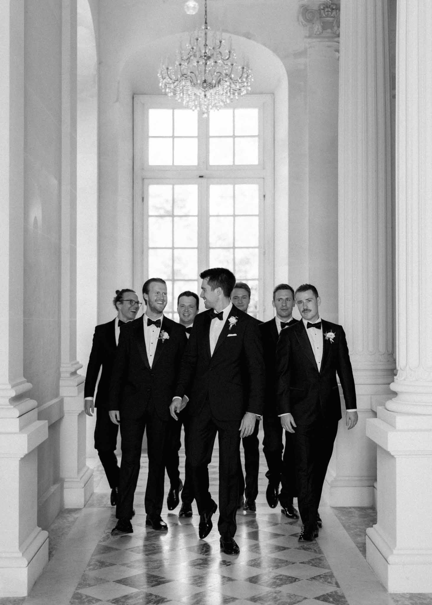 Black-and-white shot of groom and six groomsmen in black tuxedos walking down grand columned chÃ¢teau hallway