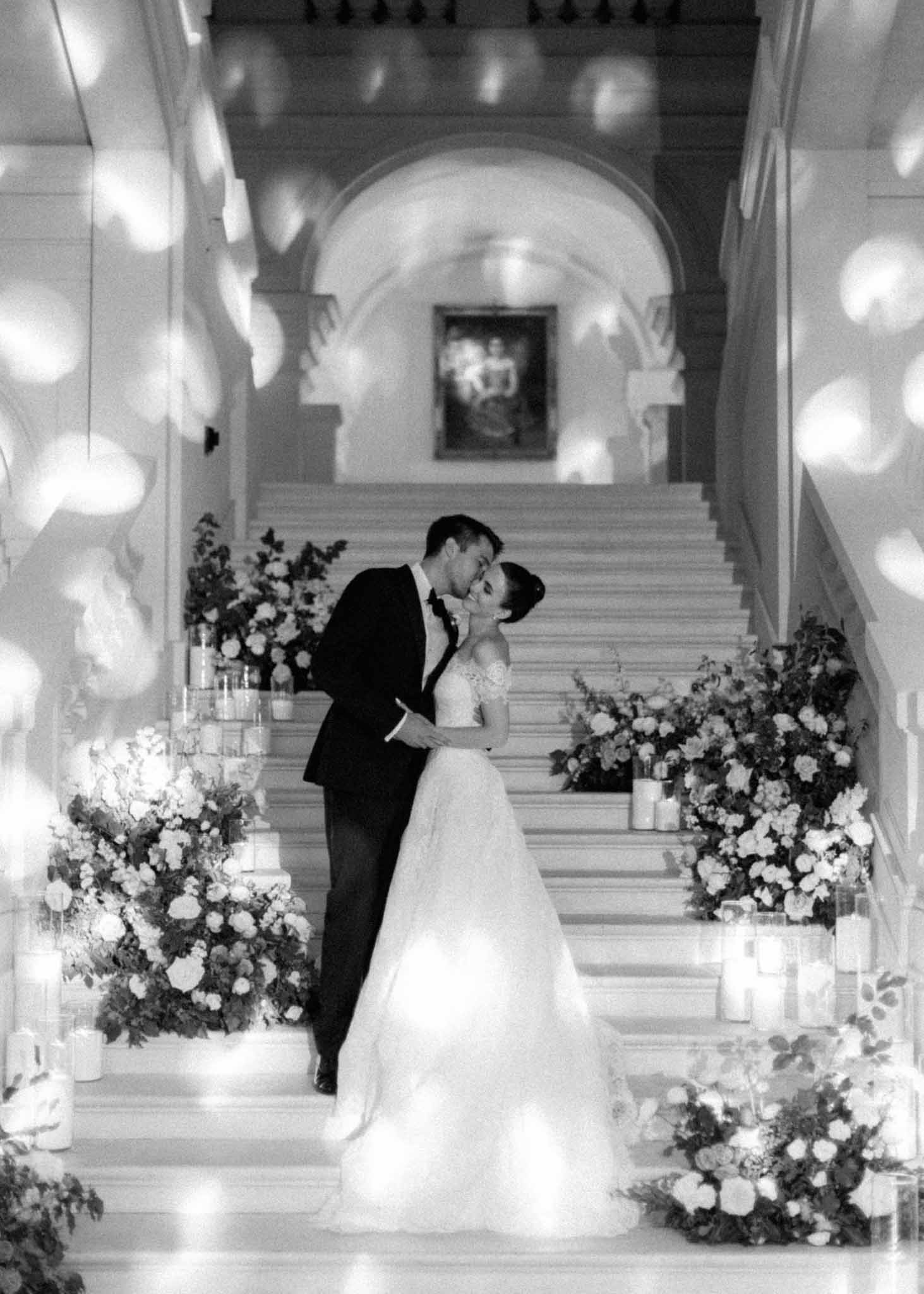 Black and white photo of bride and groom sharing first kiss in a corridor lined with white floral installations